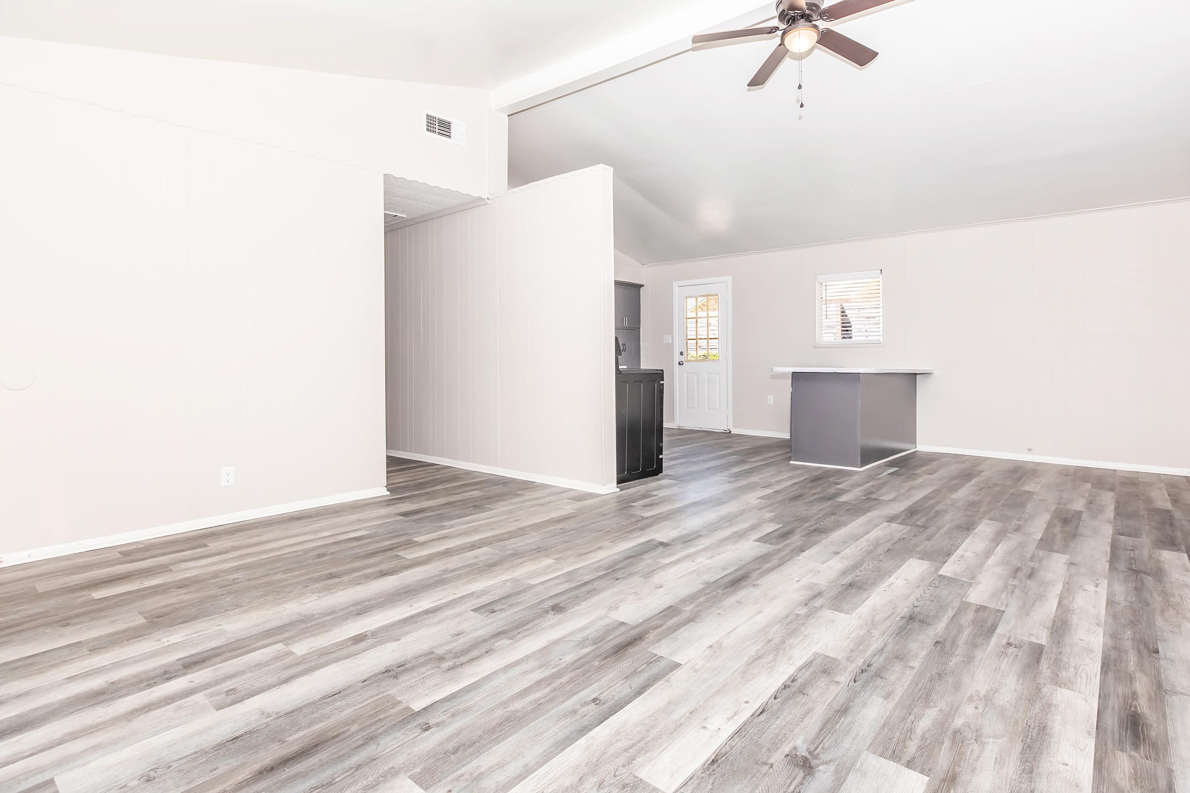 Bright, spacious room with light gray walls and a high ceiling. The floor features a modern wood-like design. To the left, there's an open doorway leading to a kitchen area. A door on the right leads outside, and a small bar counter is visible in the background. Natural light filters in through a window.