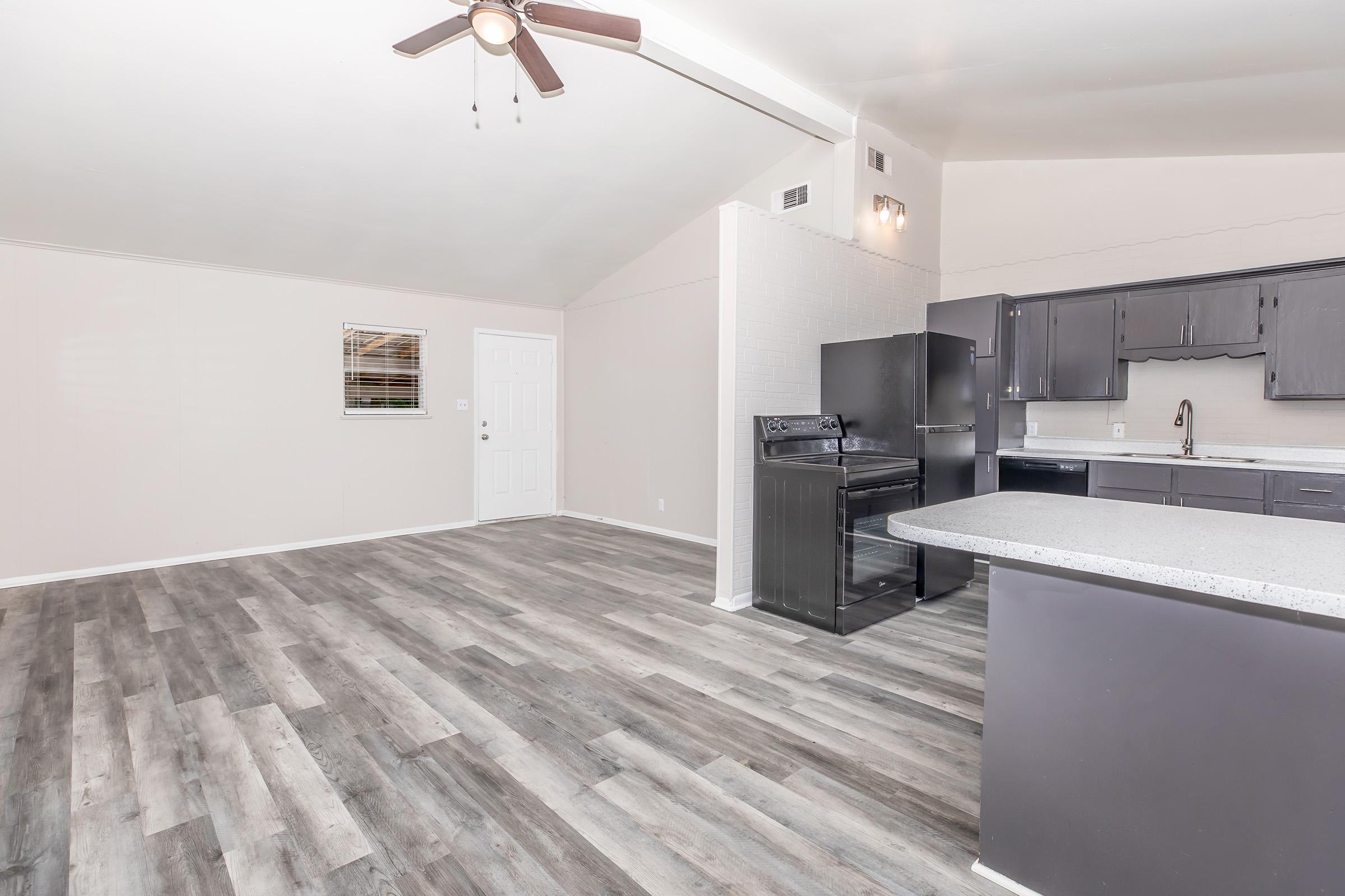 Spacious interior of a modern kitchen and living area with grey wooden flooring. Features include a ceiling fan, dark cabinetry, a black refrigerator, and an open layout that enhances natural light. The walls are painted in a light color, creating a bright and inviting atmosphere.