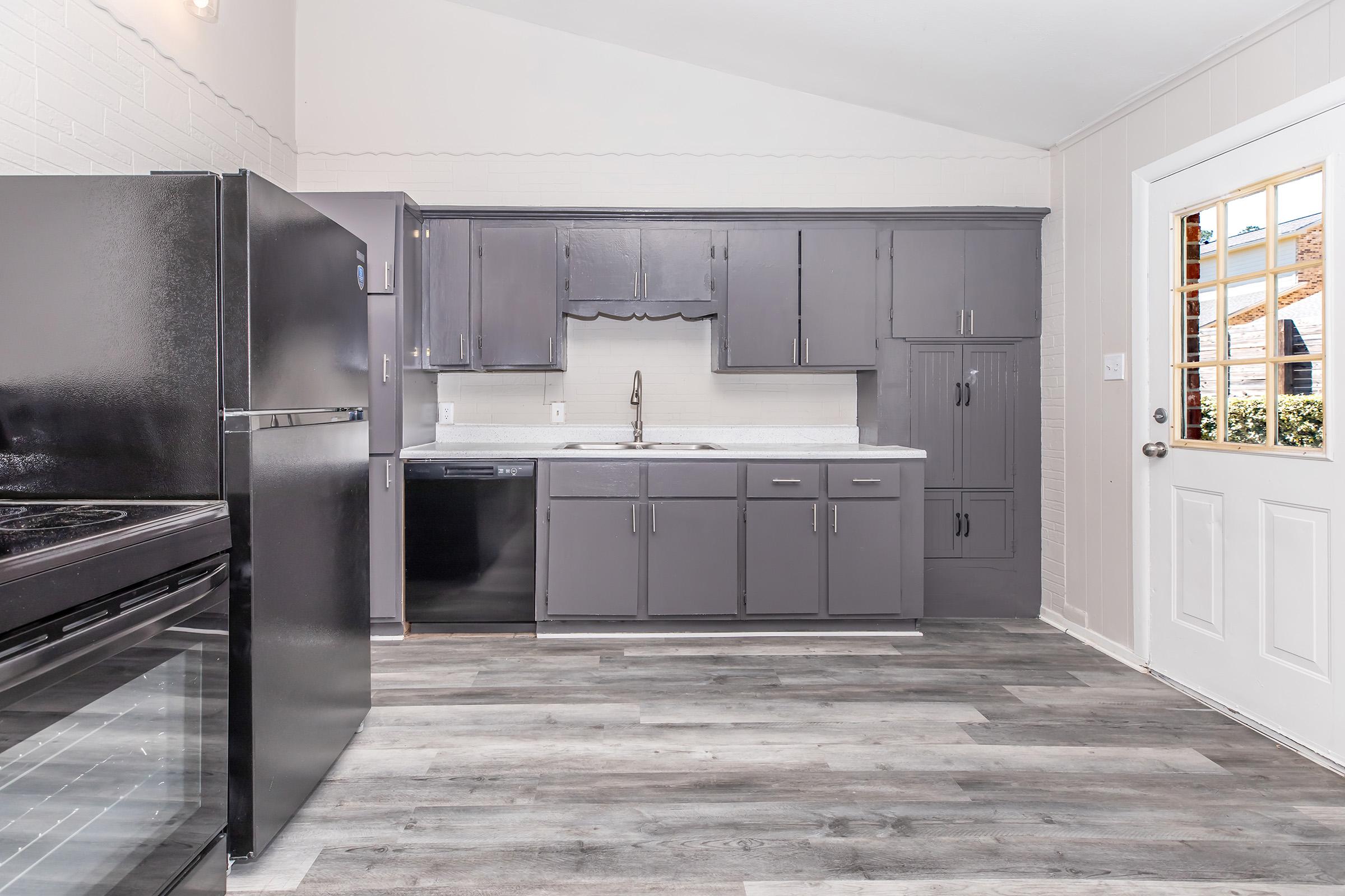 Modern kitchen featuring dark gray cabinets, white countertops, and stainless steel appliances. A black refrigerator and stove are visible, along with a sink and dishwasher. Natural light enters through a window and door, highlighting the gray wood-style flooring and clean, organized layout.