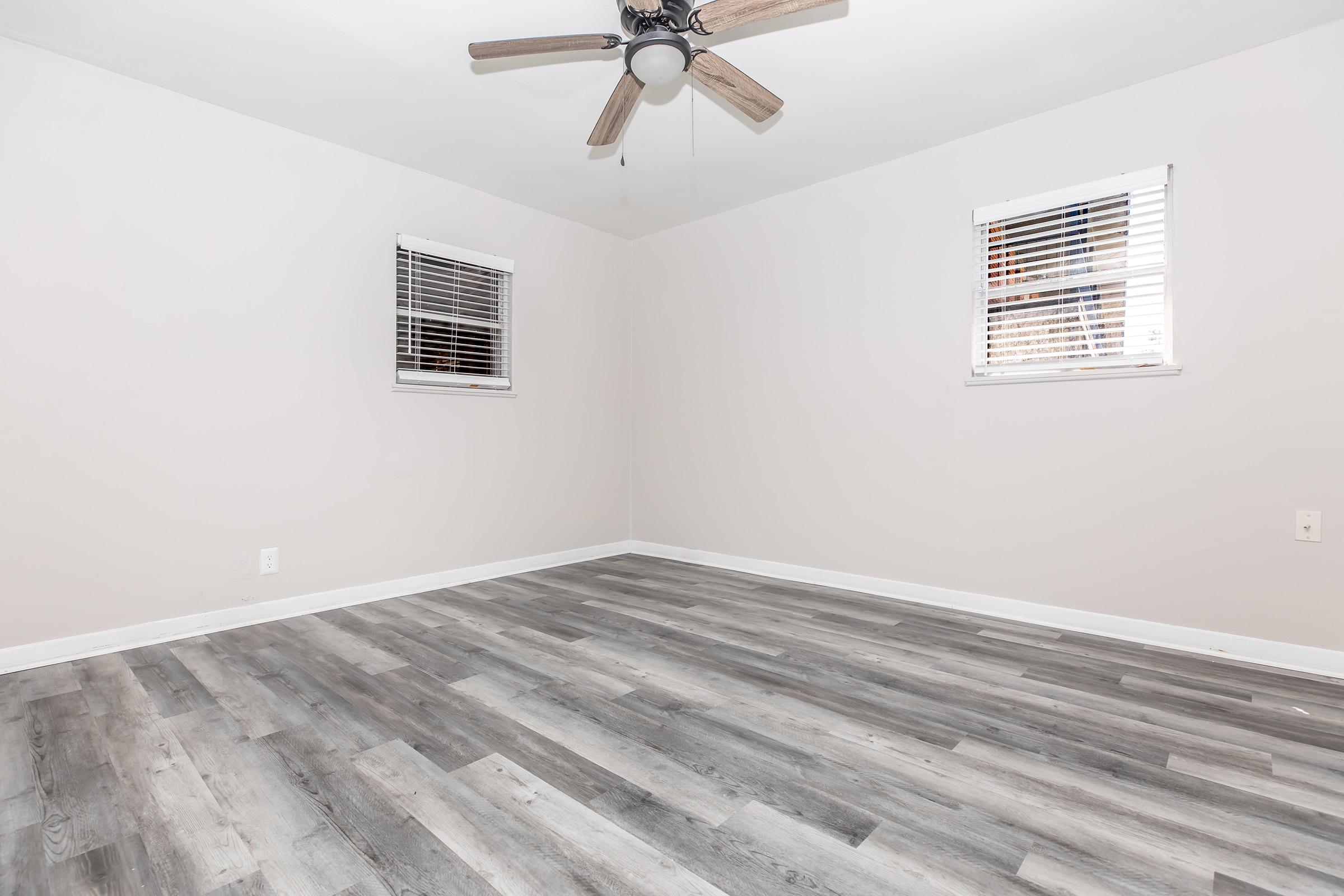 Empty room with light gray walls and gray wood-like flooring. Two small windows with white blinds on opposite walls. A ceiling fan with wooden blades hangs in the center. The space is well-lit and devoid of furniture, creating a clean and minimalistic look.