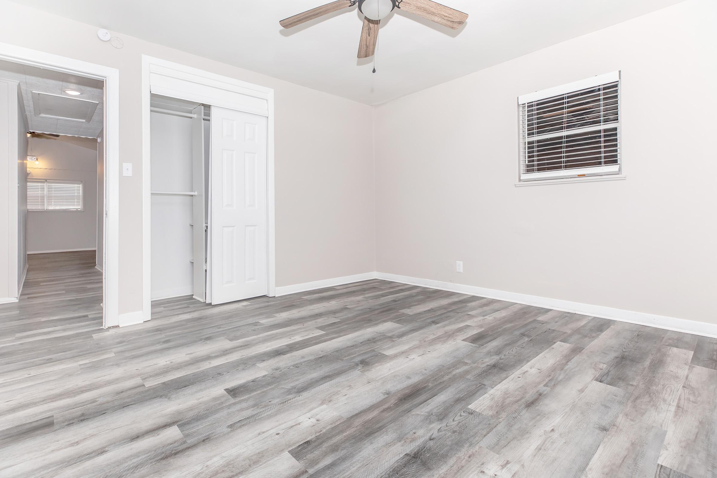 A clean, empty room with light-colored walls and gray wood-like flooring. There is a ceiling fan, a window with horizontal blinds, and an open door leading to a closet. Natural light enters through the window, creating a bright atmosphere.