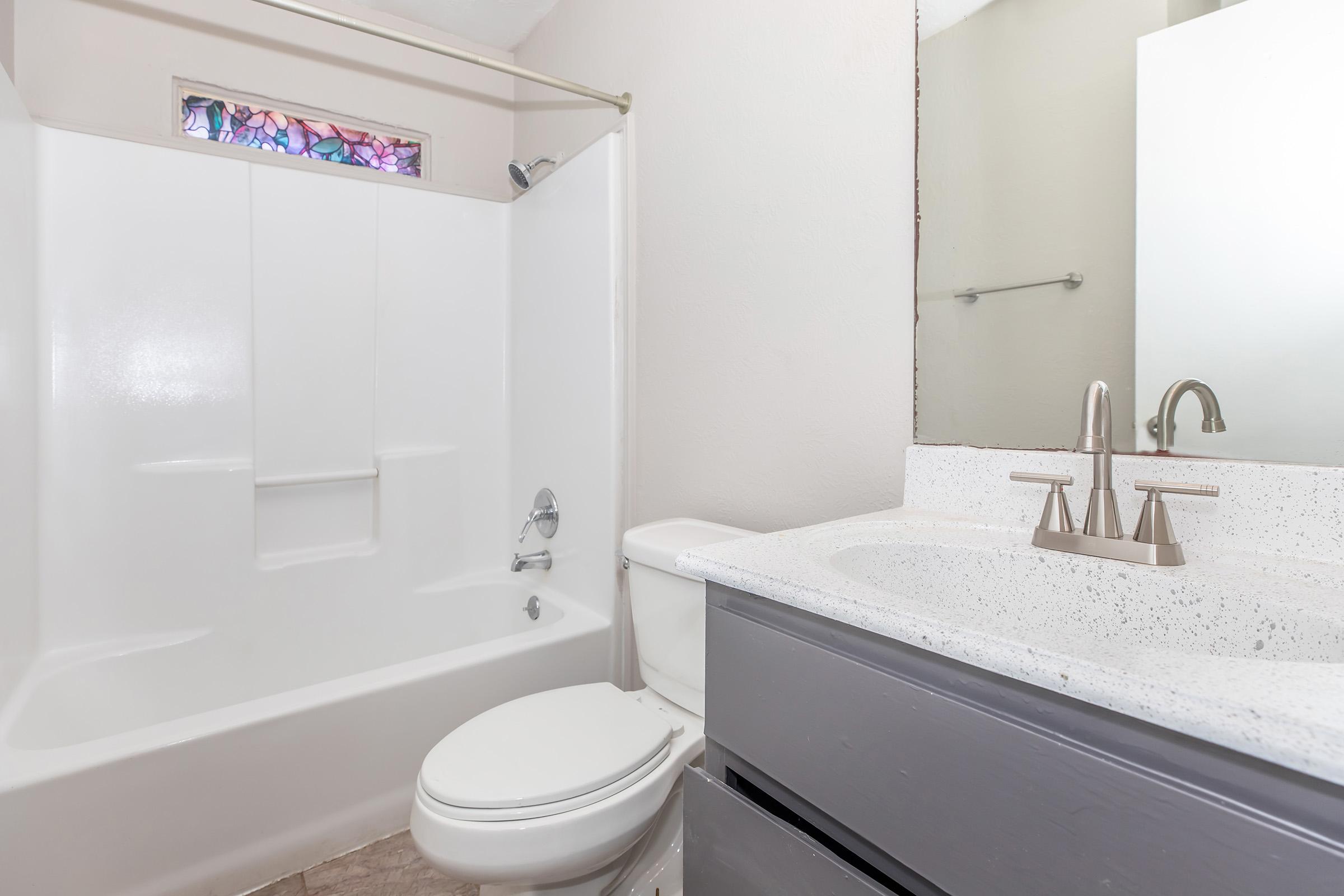 A clean, modern bathroom featuring a white bathtub/shower combination, a toilet, and a sleek vanity with a sink. The walls are painted light colors, and there is a decorative stained glass window above the tub, adding a touch of color. The flooring is a light tile, enhancing the spacious feel.