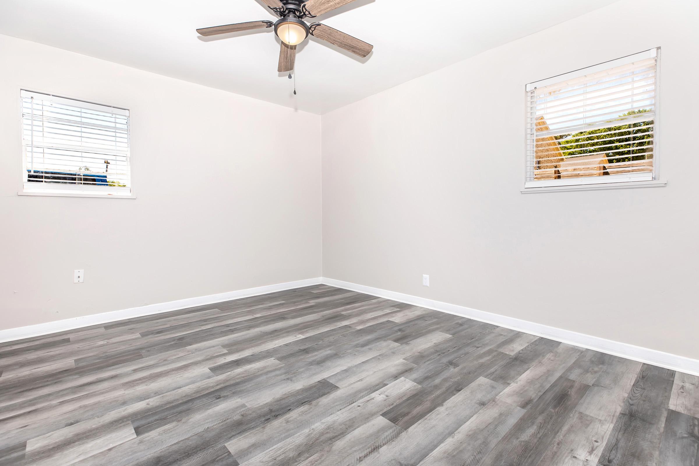 Empty room featuring a ceiling fan, light-colored walls, and two windows with blinds. The floor is covered in light gray laminate, creating a spacious and bright atmosphere.