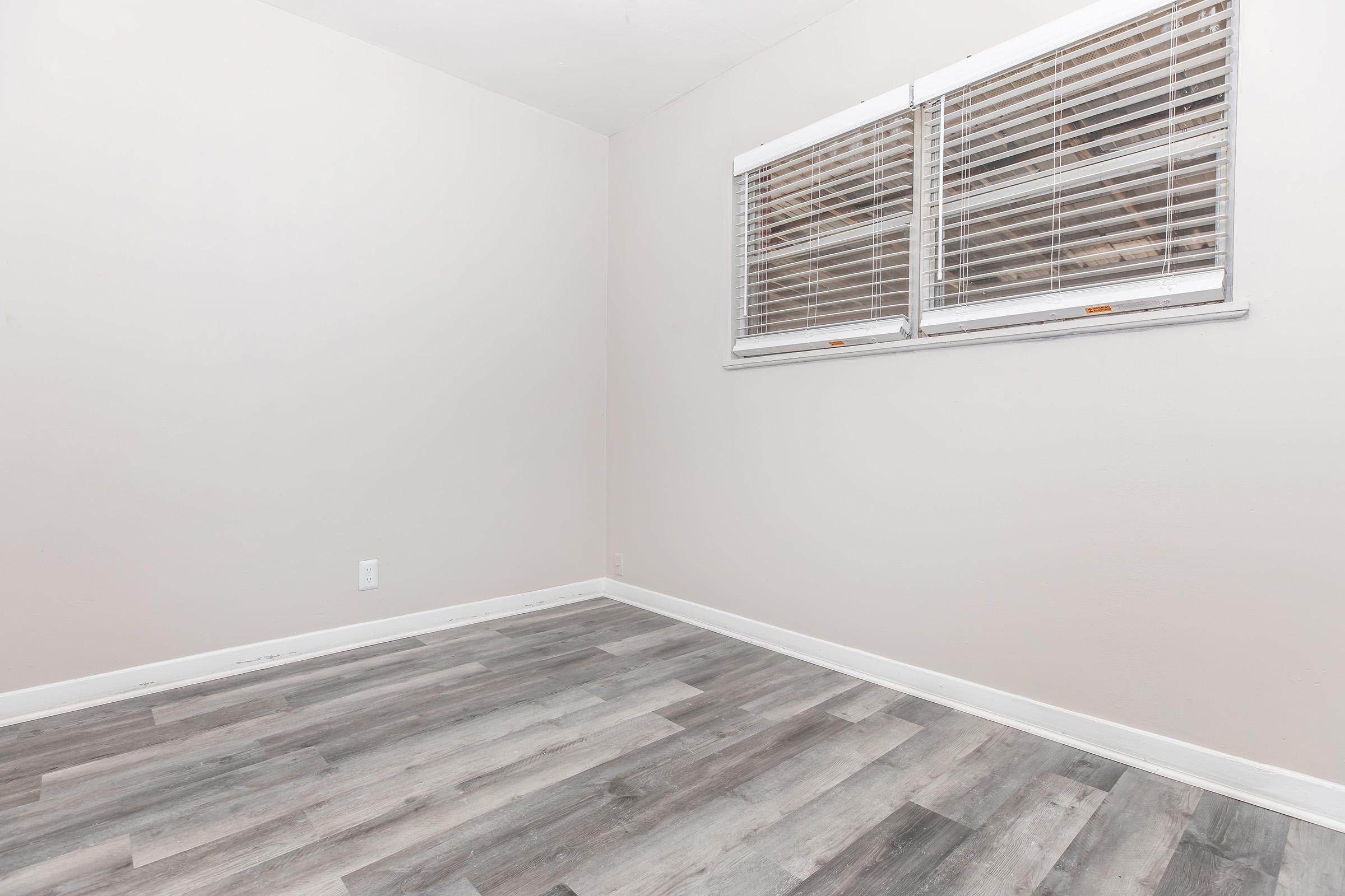 A vacant room with light beige walls and gray-green laminate flooring. A pair of white window blinds covers a window on one wall, allowing natural light to enter. The space is clean and unfurnished, creating a bright and simple aesthetic.