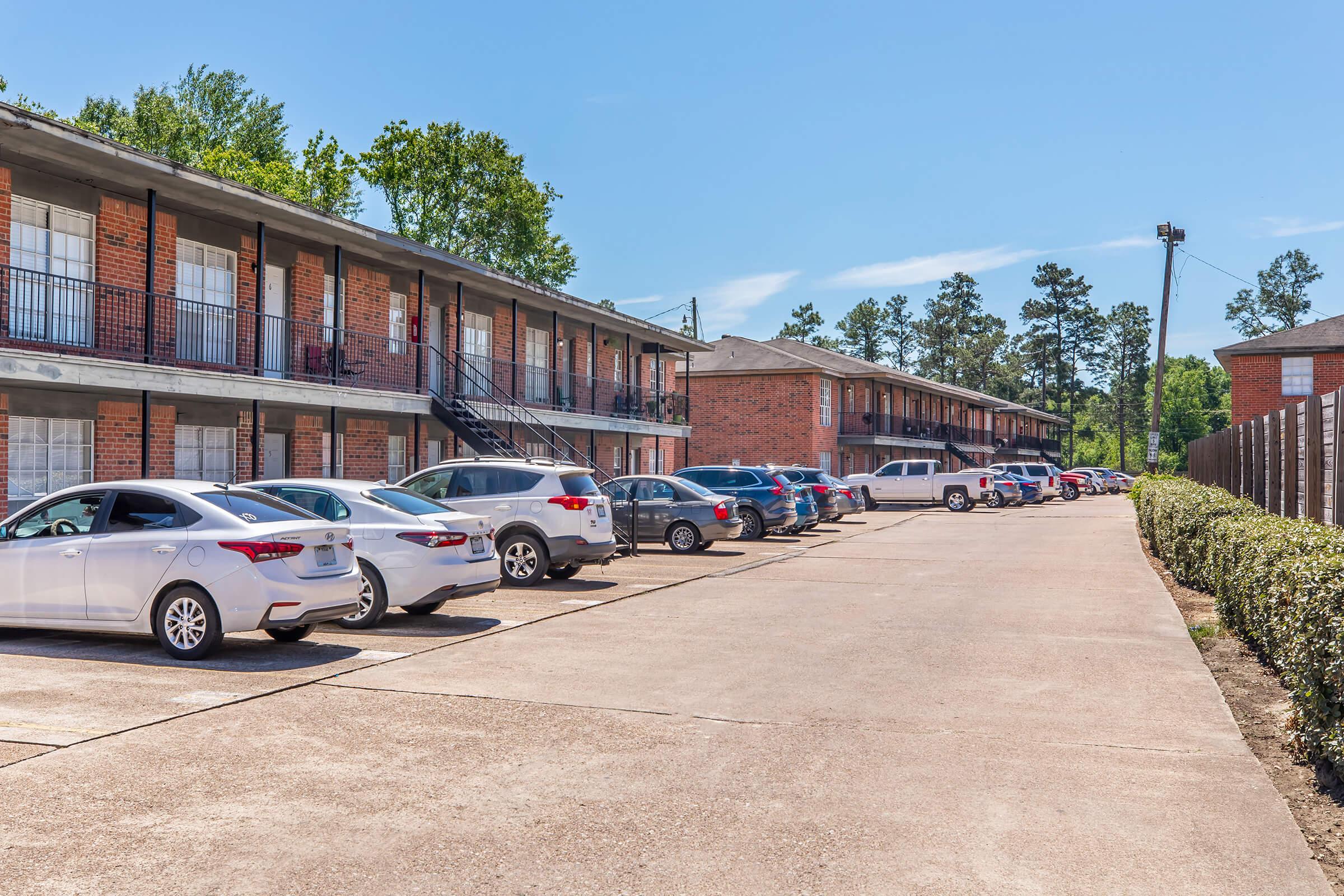 View of a residential apartment complex featuring two-story brick buildings with balconies. Several cars are parked along a paved driveway, with landscaped greenery and trees in the background under a clear blue sky.