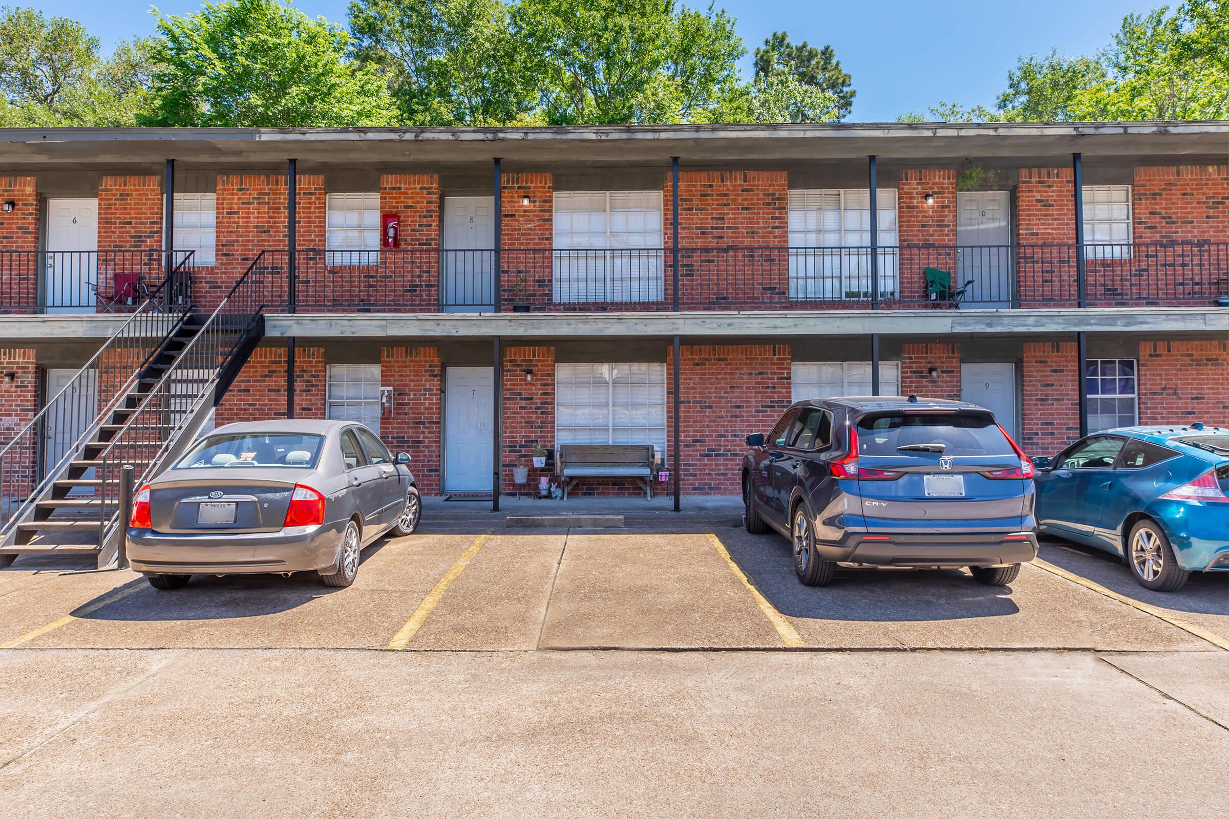 A view of a two-story brick apartment building with several parking spaces in front. The building features multiple doors and balconies, surrounded by greenery. Three parked cars are visible in the foreground, with one grey sedan, one dark SUV, and one blue vehicle. The setting appears sunny and well-maintained.
