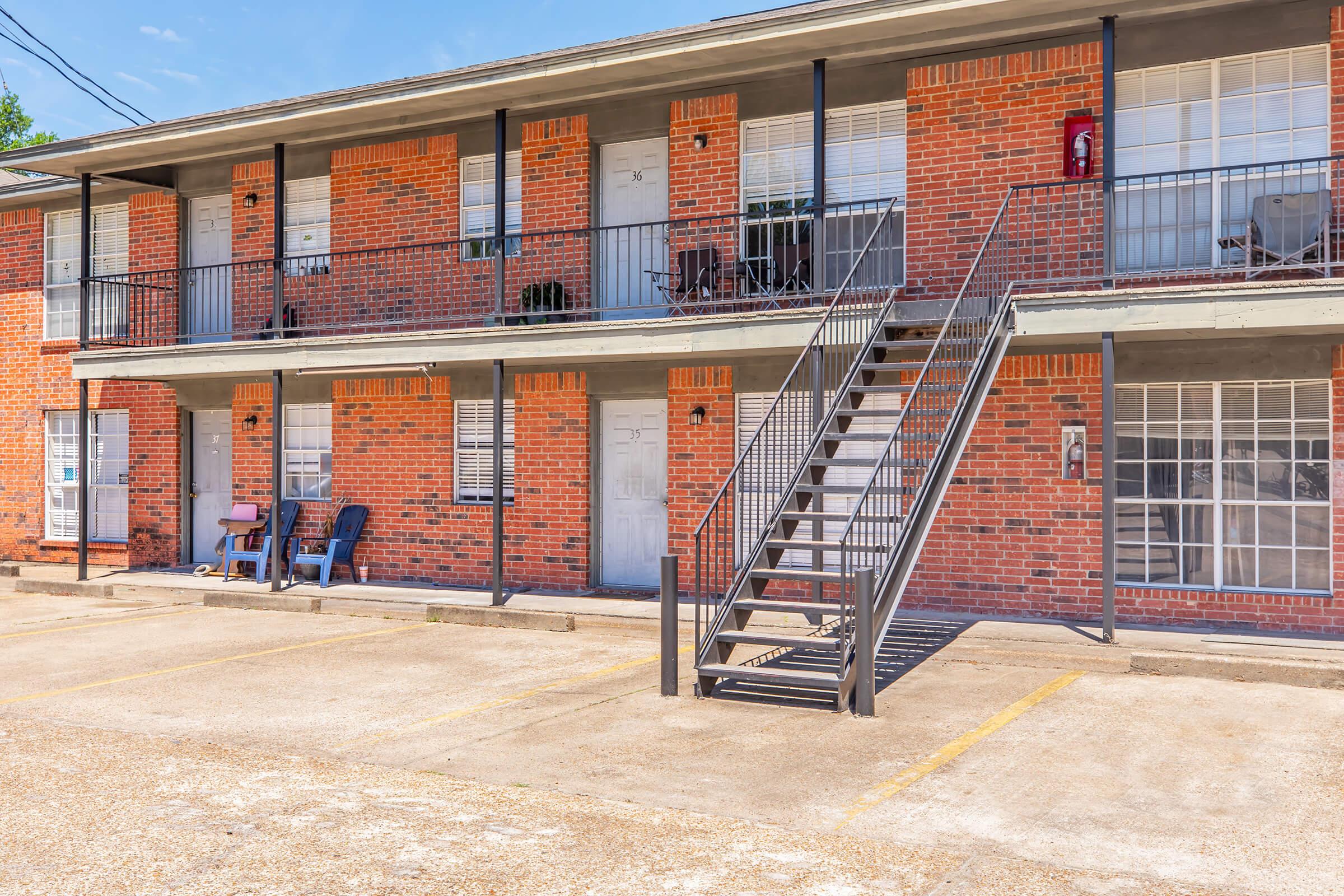 Two-story brick apartment building with a staircase leading to the upper level. Ground floor features several doors, with chairs placed outside. The parking lot is visible in front, marked with yellow lines, and the sky is clear blue. A fire extinguisher is mounted on the wall near the entrance.