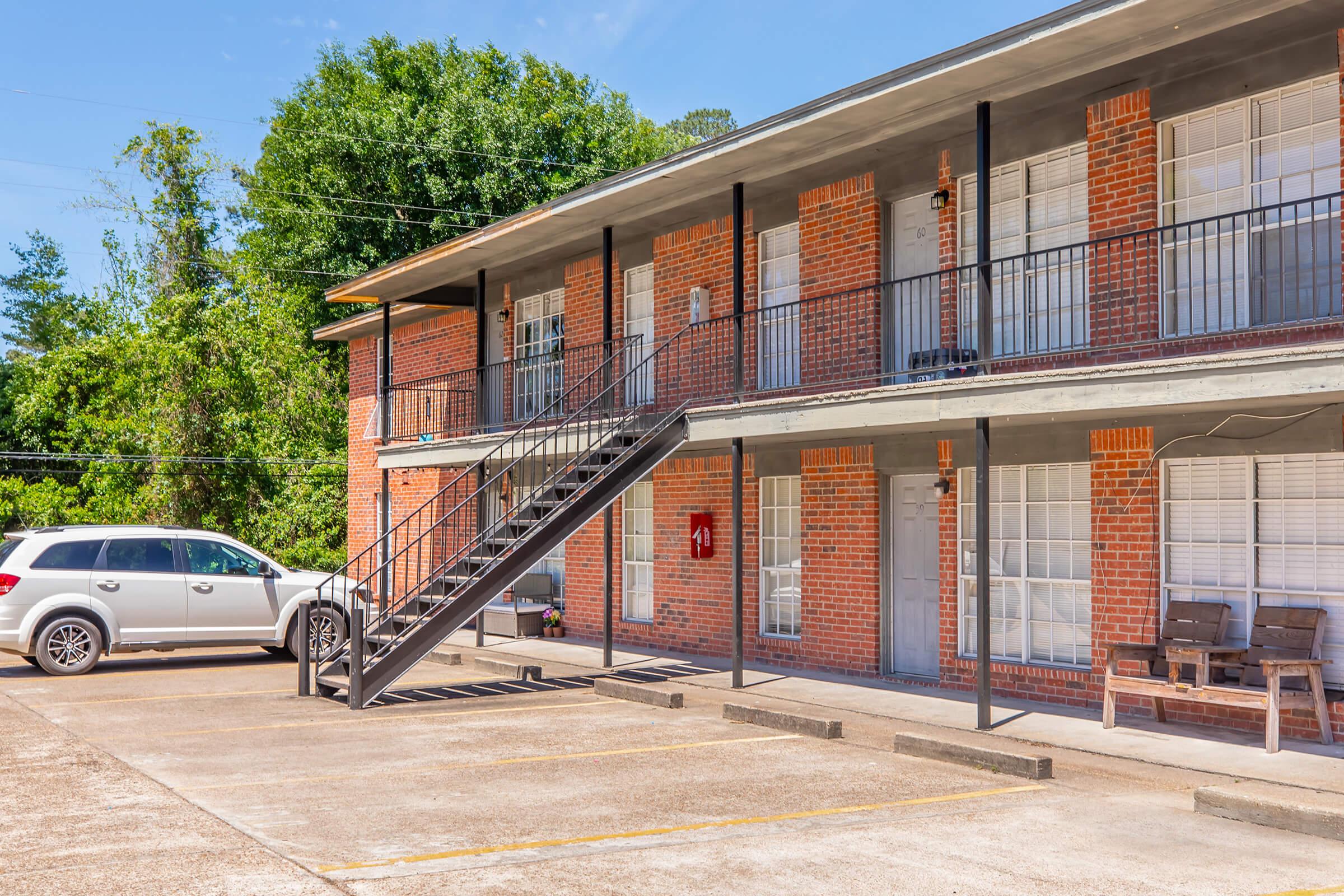 A two-story brick apartment building featuring a staircase on the left, with white railings and dark gray trim. Several windows are visible, some with blinds. A silver SUV is parked in front, and there are grassy trees in the background under a clear blue sky.