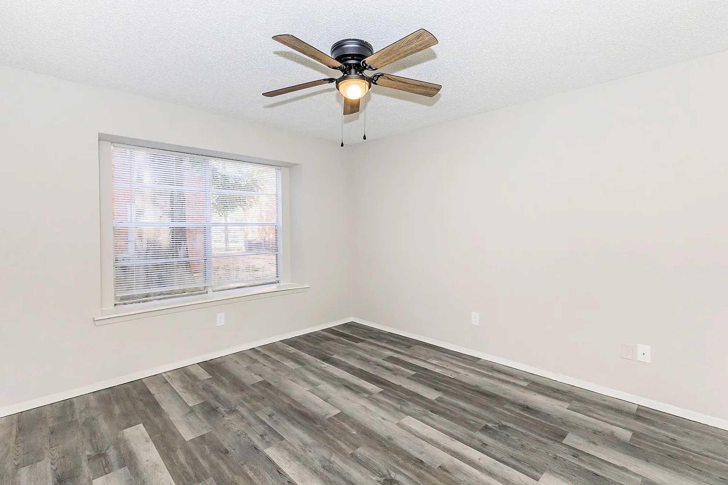 A spacious, empty room with light gray walls and a ceiling fan. The floor has a modern wood-like design. A large window allows natural light to enter, illuminating the space. The overall ambiance is clean and inviting, perfect for personalization or setting up furnishings.