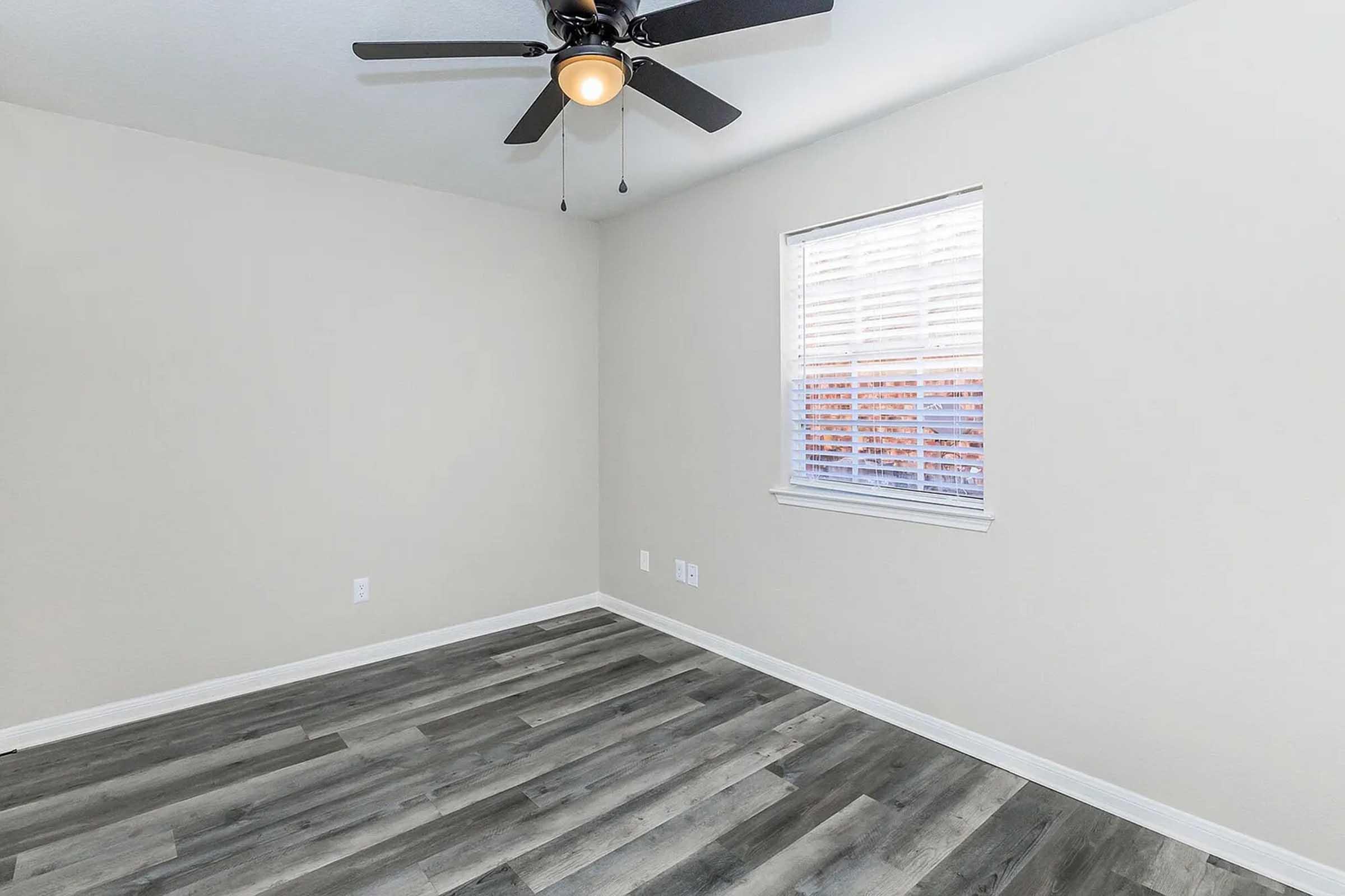 A simple, empty room featuring light-colored walls, a ceiling fan with a light fixture, and white plantation-style window with blinds. The floor is composed of gray laminate planks, and there's minimal furniture or decor present, emphasizing the room's spaciousness and simplicity.