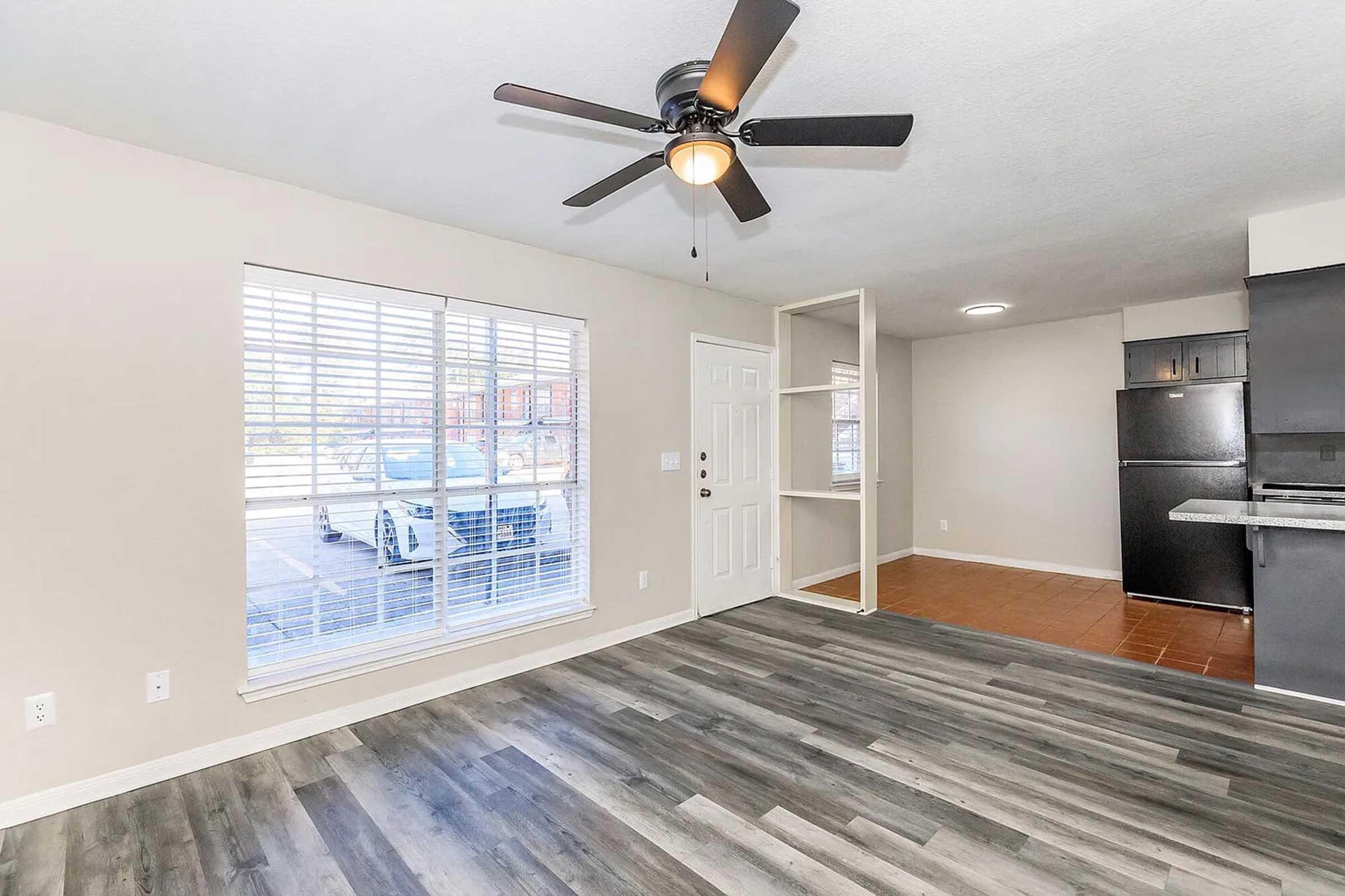 A well-lit living space featuring a ceiling fan, large window with blinds, and laminate flooring. A white door leads outside, while a small shelf is visible on the right. The kitchen area includes a black refrigerator and is in an open layout. Natural light fills the room, creating a welcoming atmosphere.