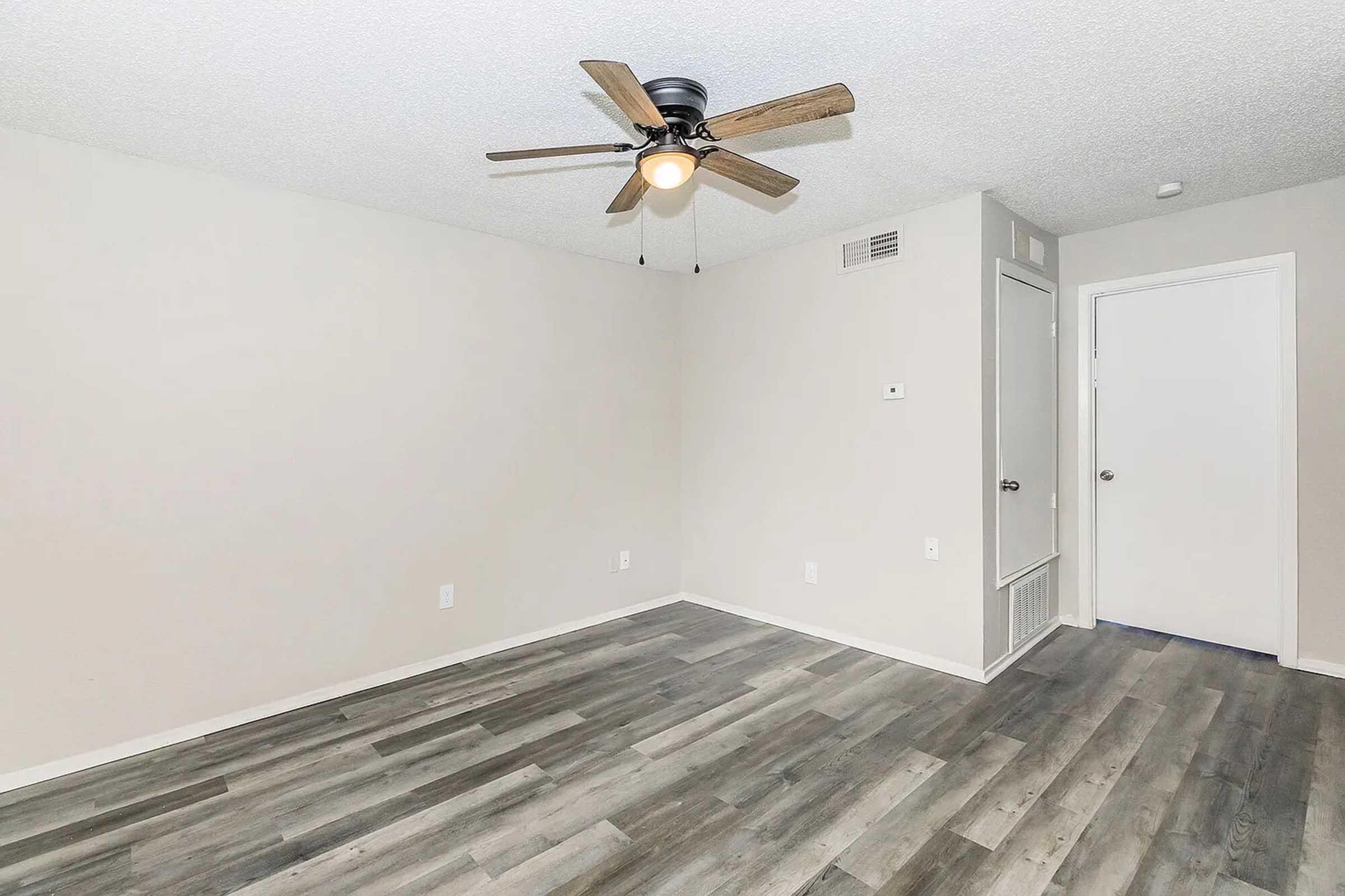 A spacious room with light-colored walls and a ceiling fan. The floor is covered in light wood-like laminate. There is a door on the right side leading to an adjacent area, and a small wall vent. The room is empty, creating a bright and open feel.