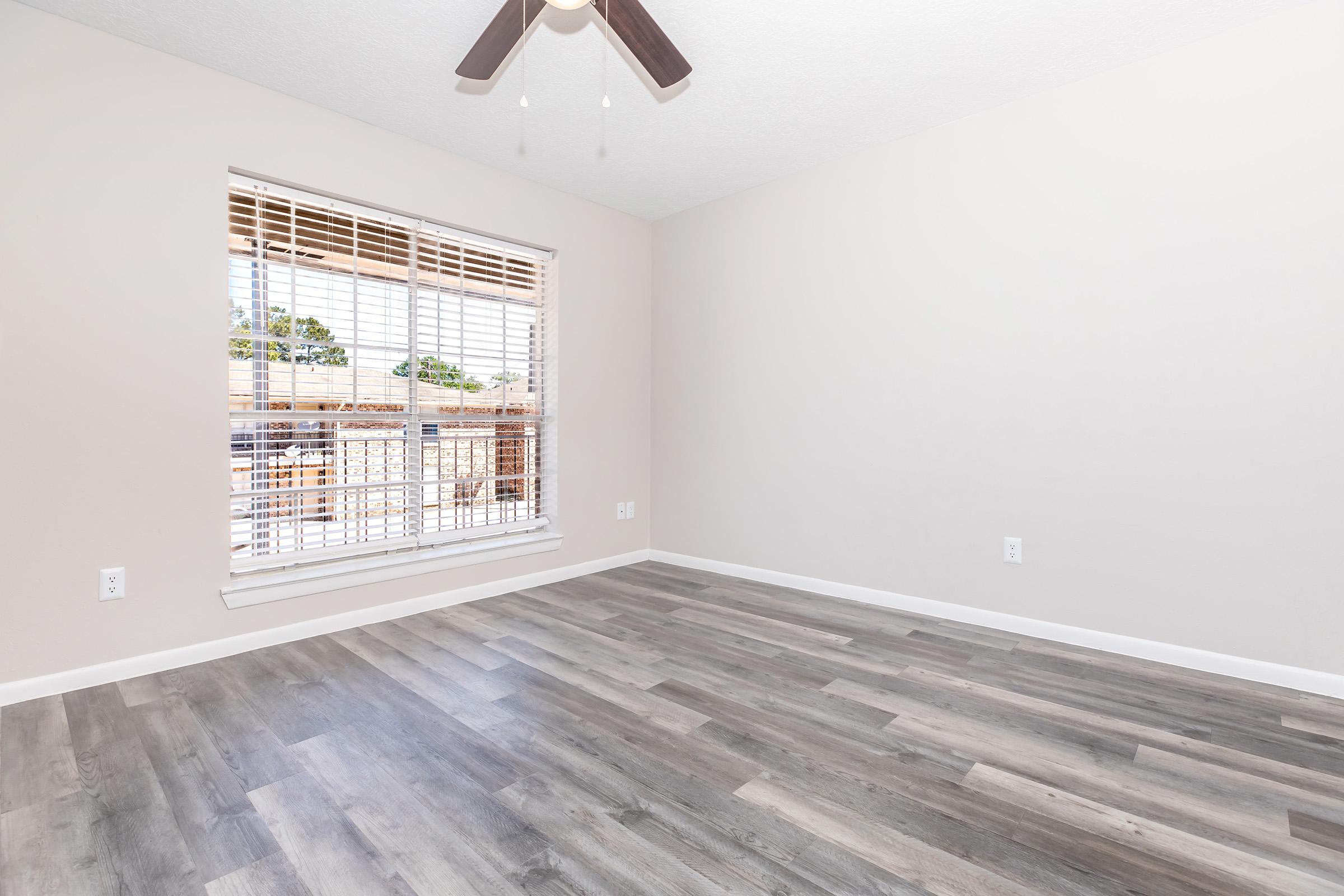 A bright, empty room featuring light gray walls and a large window with white blinds. The floor is covered in light wood-look laminate. A ceiling fan with wooden blades hangs from the ceiling, complementing the neutral decor. Natural light spills in, creating an airy atmosphere.