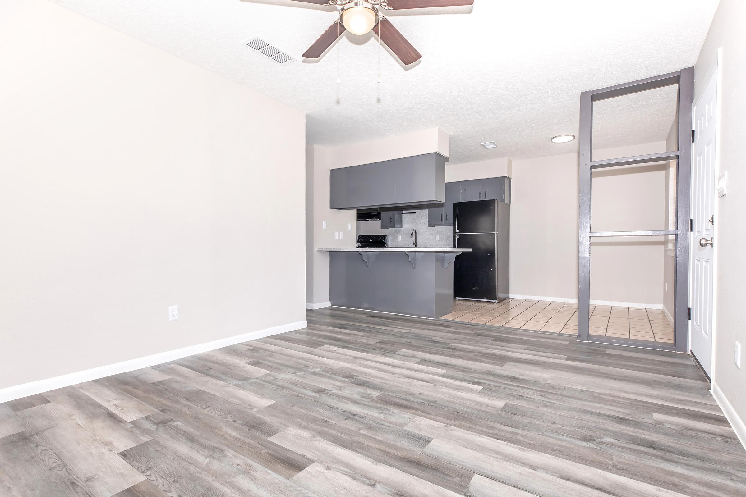 A spacious room featuring light-colored walls and a ceiling fan, with wooden flooring. To the left, there's a modern kitchen area with dark cabinets and appliances. A glass partition provides separation from another area, enhancing the open feel of the space. Bright and inviting atmosphere.