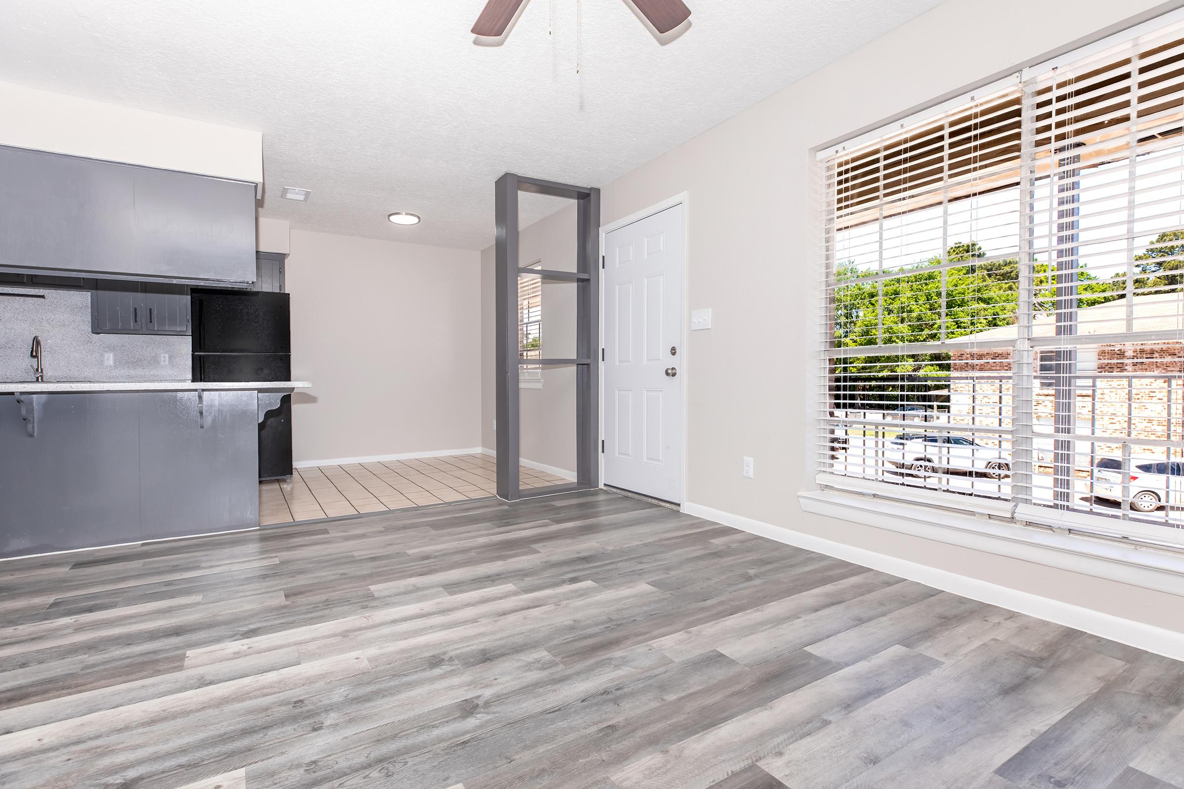 A spacious, well-lit room featuring a modern kitchen area with black cabinets and appliances. The flooring is a stylish gray laminate, and there's a window with blinds that offers a view of the outside. The room has a ceiling fan and a white door leading to another space, creating an open and inviting atmosphere.