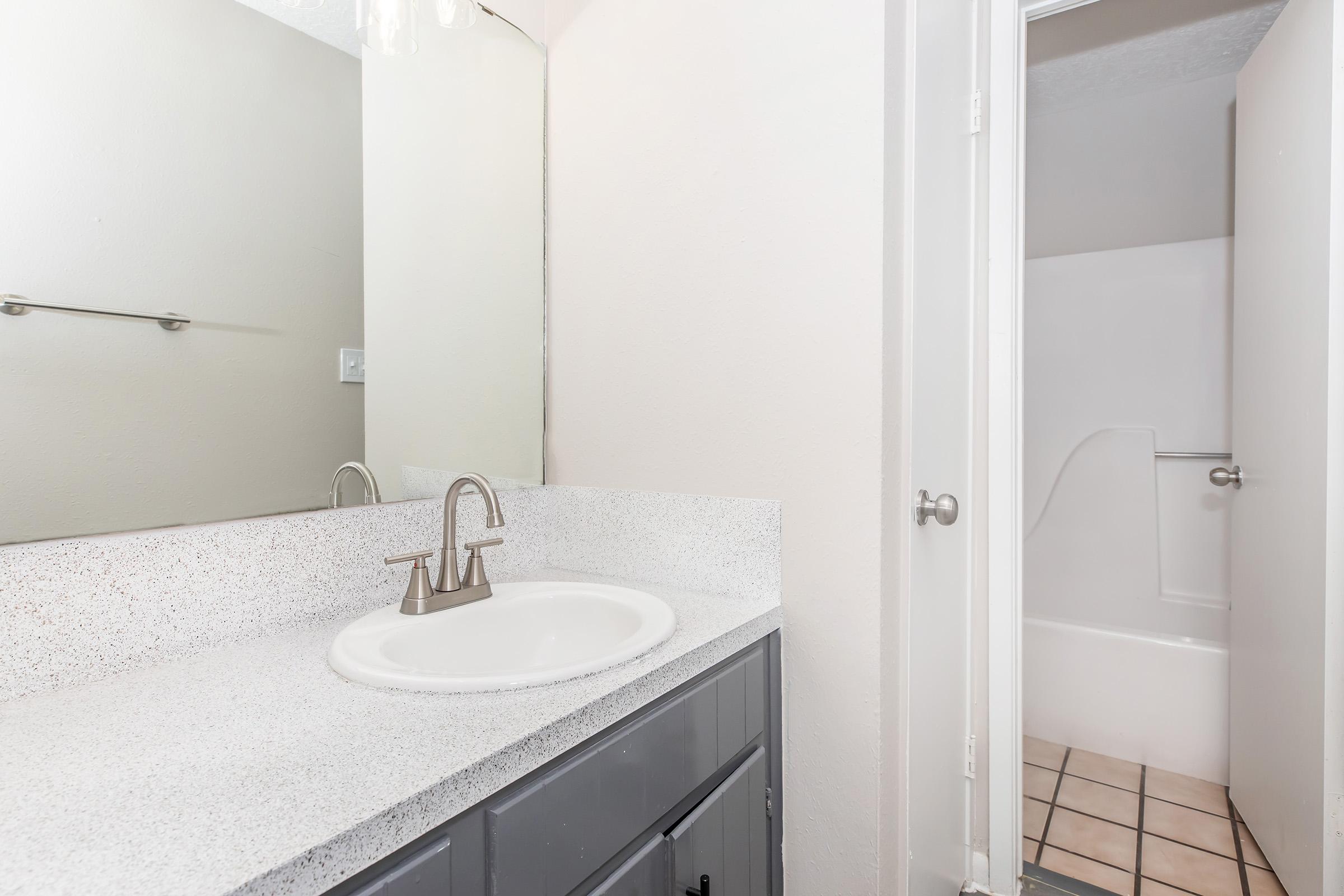 A clean, modern bathroom featuring a gray vanity with a round sink and a sleek faucet. The walls are painted a light color, complementing the granite-like countertop. A full bathtub is visible through an open door, and the tiled floor adds a stylish touch to the space.