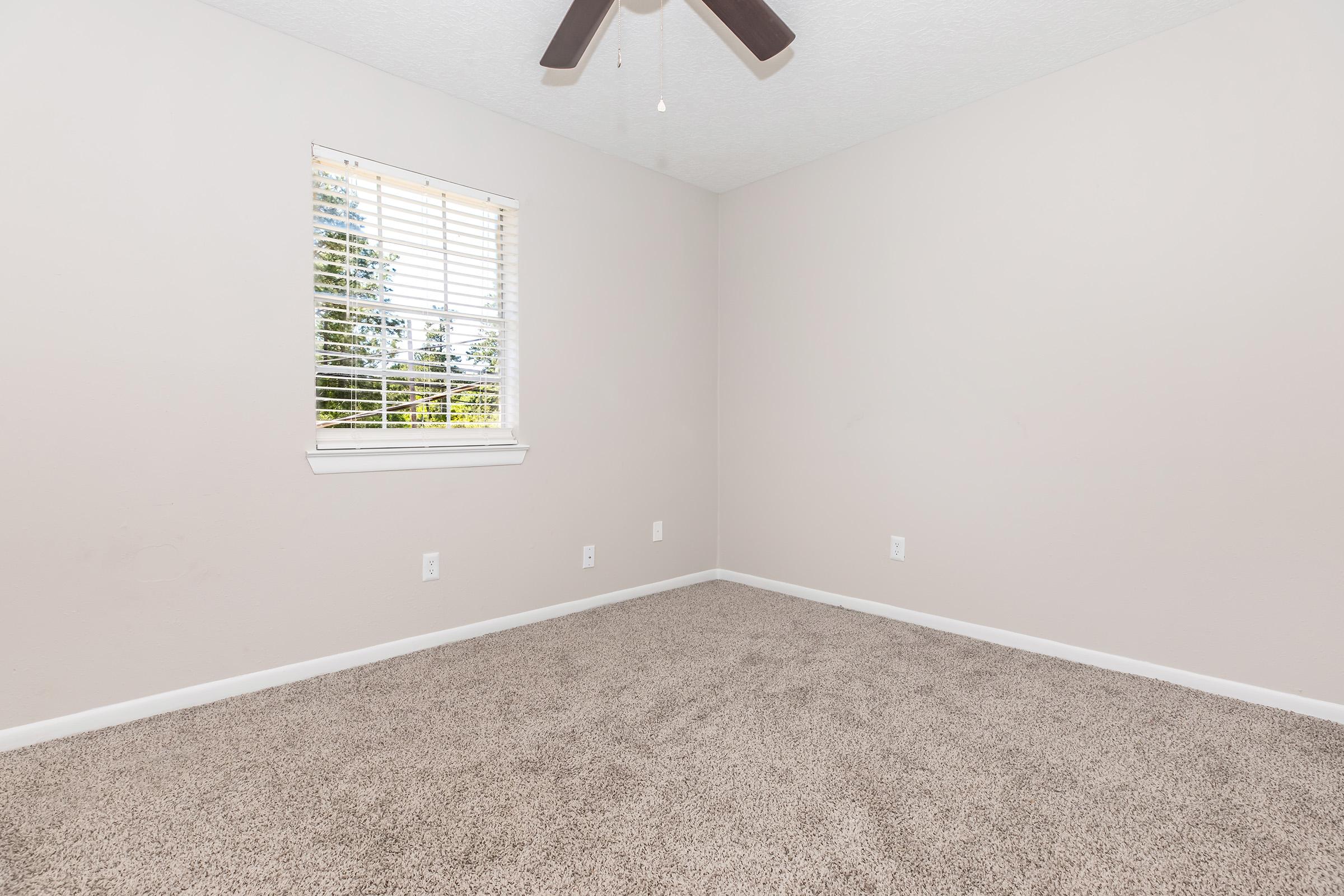 Empty room with light beige walls and tan carpet. A window with blinds allows natural light to enter, while a ceiling fan hangs above. There are no furnishings or decorations, creating a spacious and minimalist atmosphere.