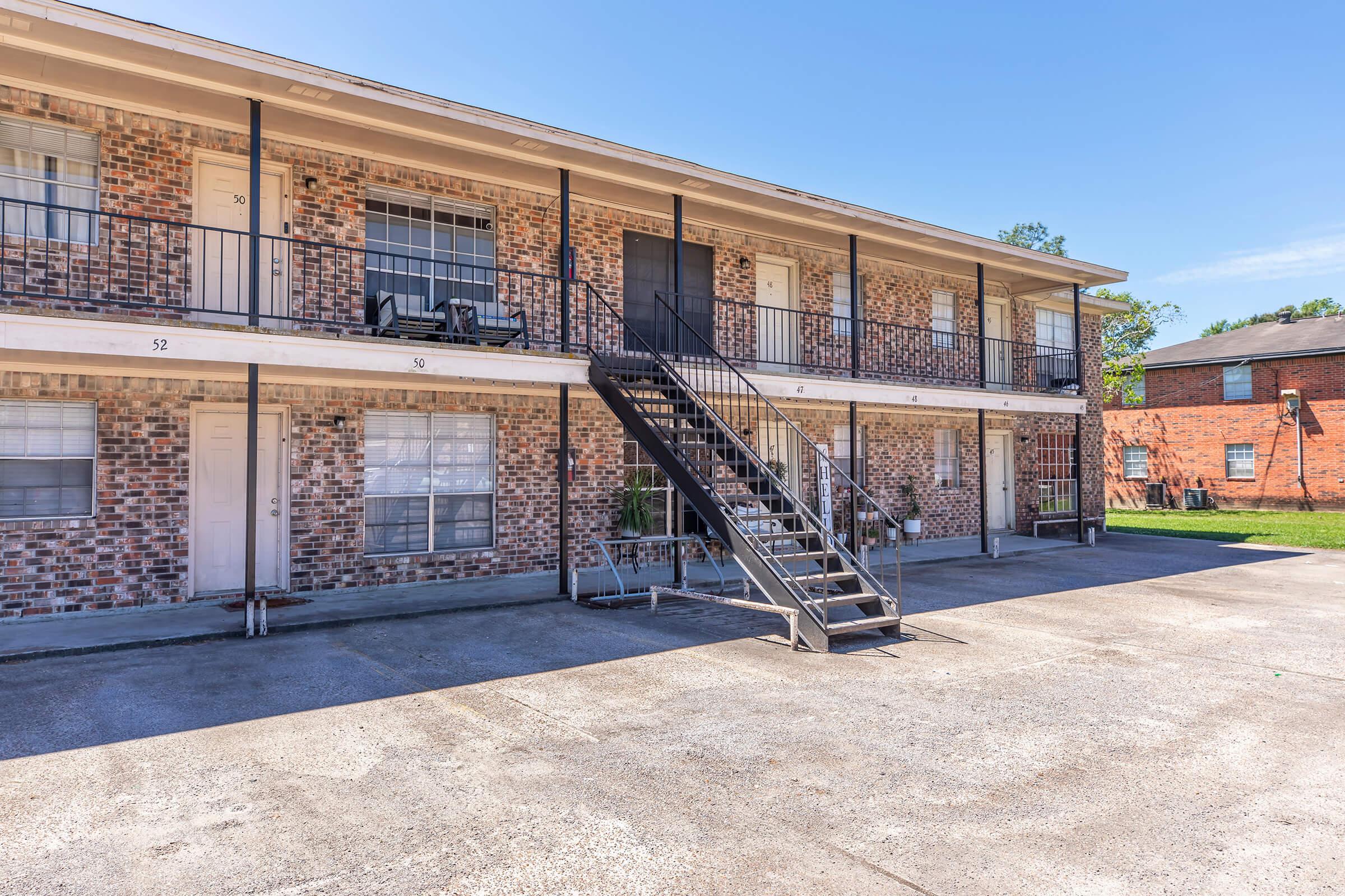 A two-story apartment building with brick exterior and balconies. The ground floor features several white doors, with stairs leading up to the second level. The area in front has a concrete parking space, and grassy areas are visible in the background under a clear blue sky.