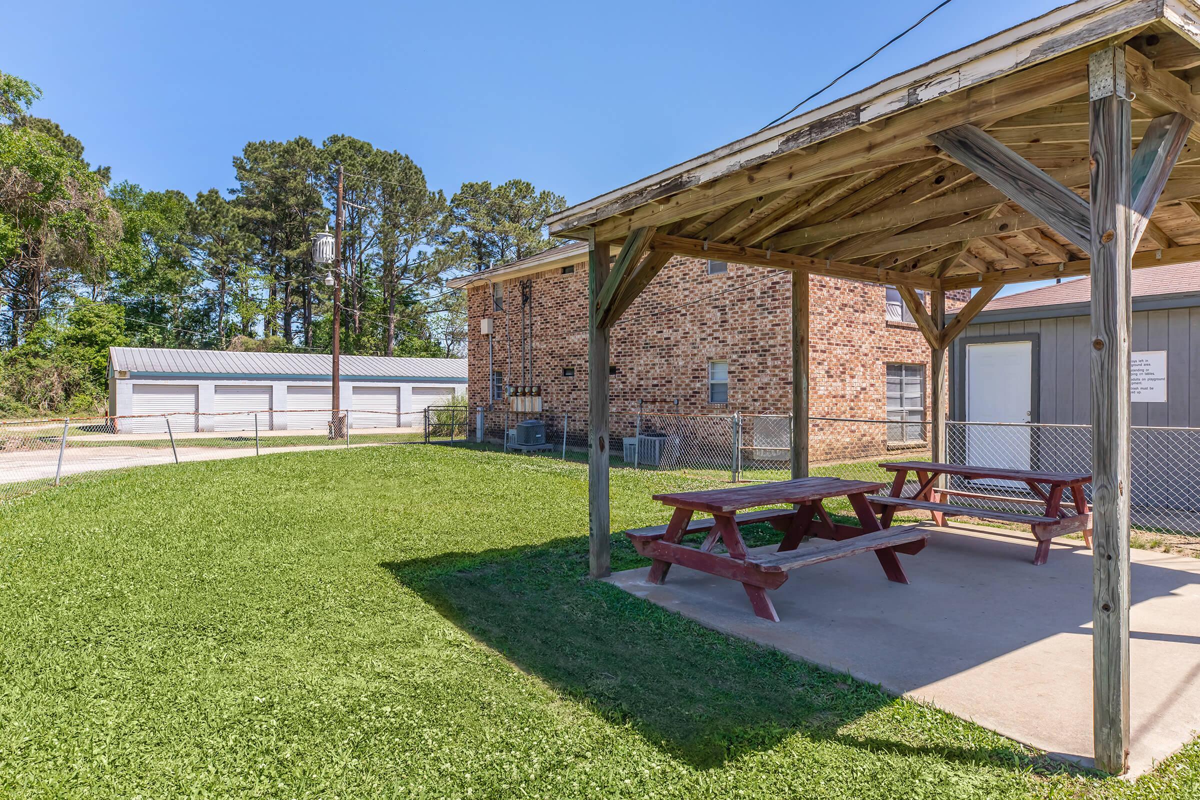 A shaded picnic area with wooden tables under a roof, surrounded by green grass. In the background, there are storage units and a brick building. The scene features clear blue skies and a few trees, providing a tranquil outdoor setting.