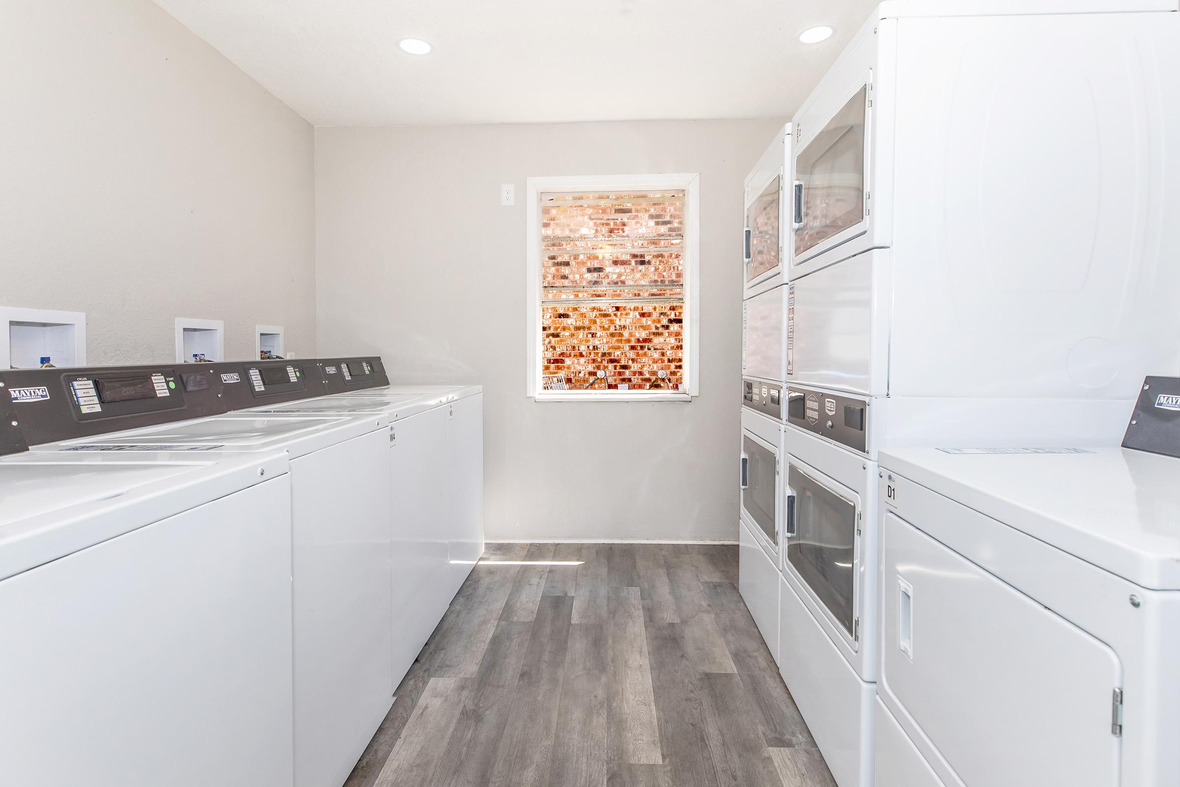 A bright, clean laundry room featuring several white washing machines and dryers aligned against the walls. A window with a textured glass design allows natural light to fill the space, highlighting the modern, minimalist design and sleek appliances. The floor is covered with light wood-like laminate.