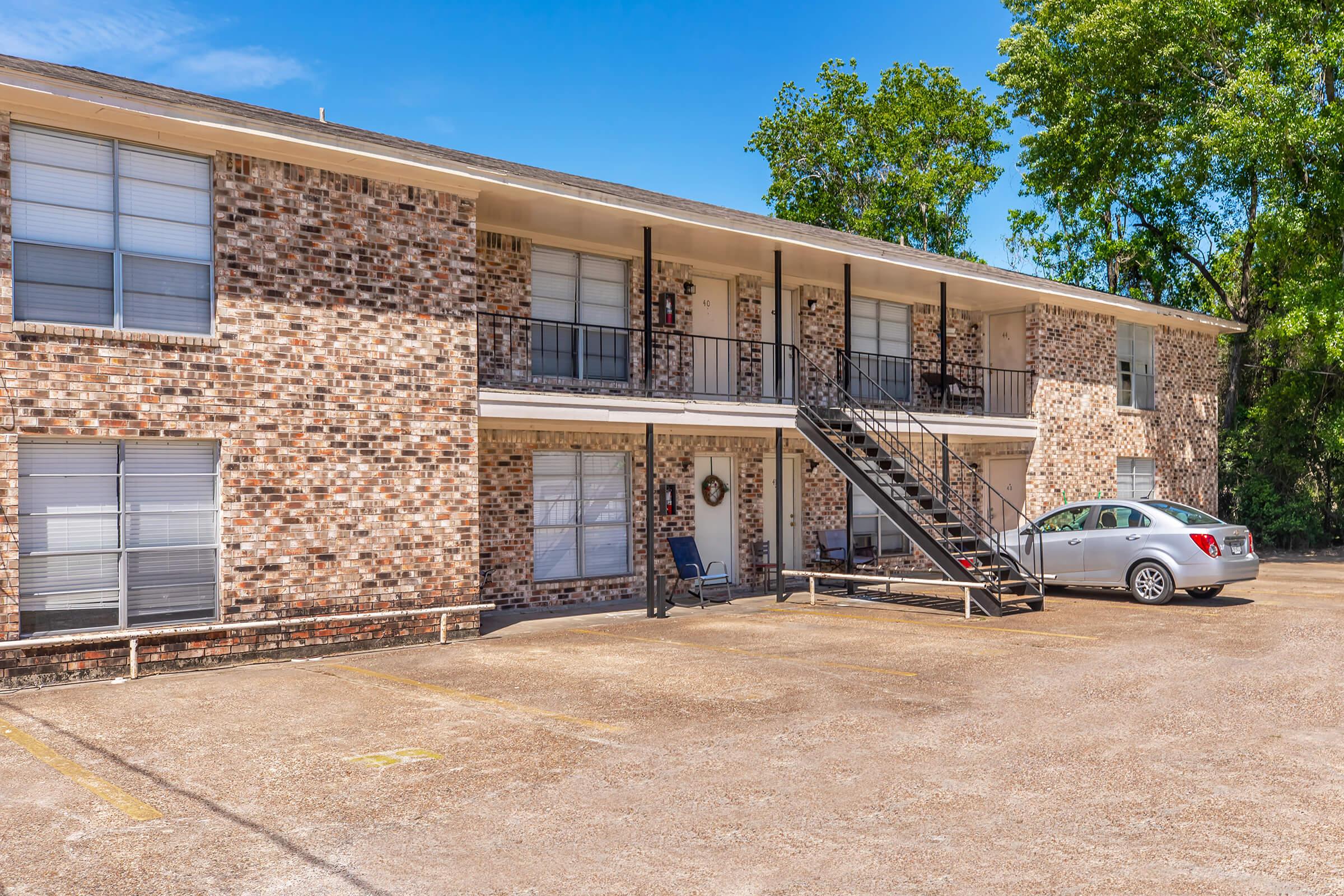Two-story brick apartment building with a staircase leading to the second floor. The ground level features several windows and a small porch area. A light-colored car is parked in the parking lot in front of the building, which is surrounded by trees and a blue sky.