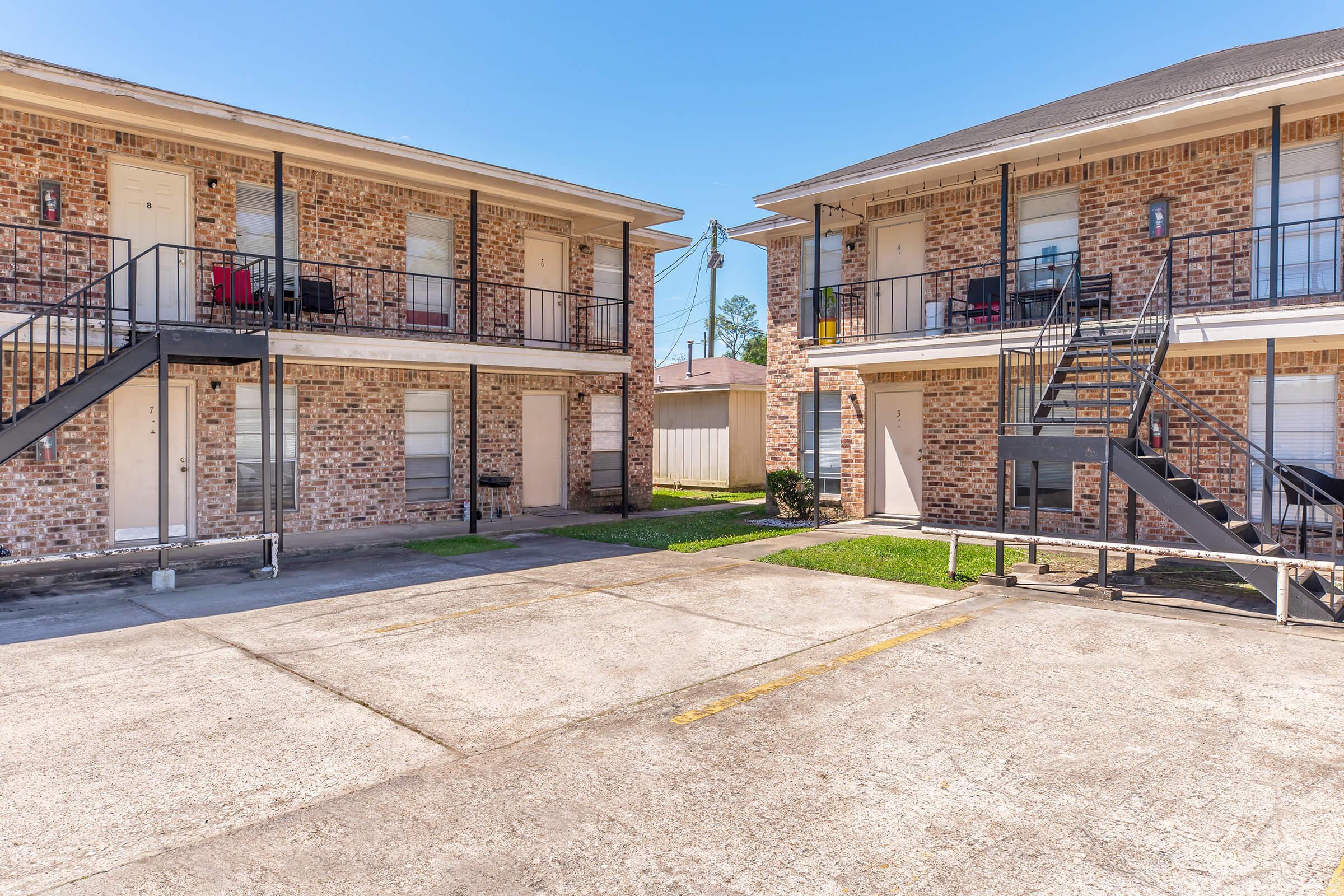 A view of a two-story brick apartment complex with outdoor staircases. The courtyard area is paved, and there are grassy patches. Several apartment doors are visible, some with porch furniture. Bright blue sky overhead adds to the sunny atmosphere.