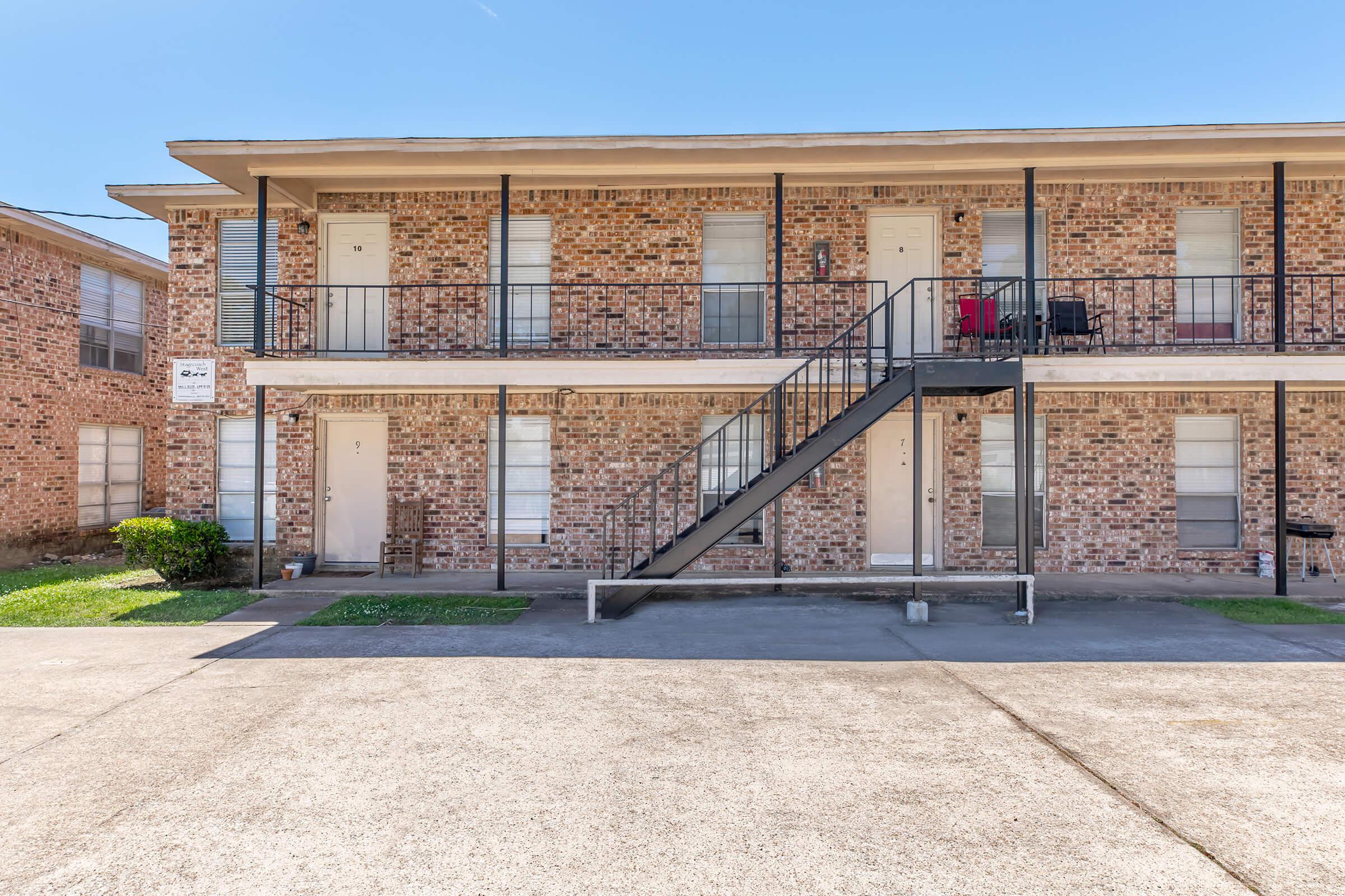 Two-story brick apartment building with multiple units. The ground floor has three doorways, each with windows beside them. A black metal staircase leads to the second floor, which has additional doorways. The area features a small patch of grass and a clear blue sky above.