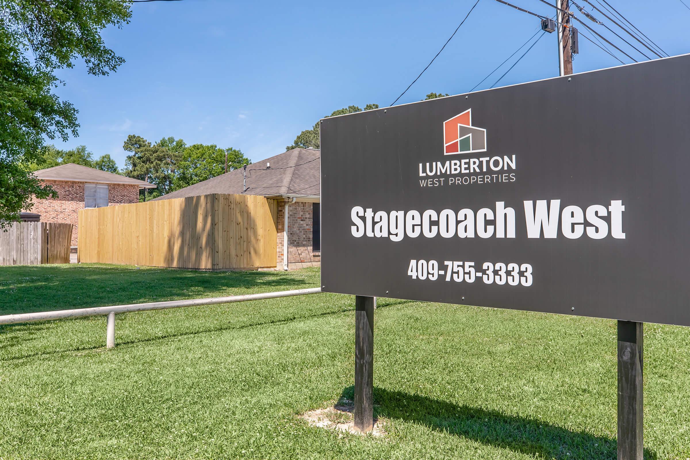 Sign for Stagecoach West, a property managed by Lumberton West Properties. The sign includes the contact number 409-755-3333. In the background, there are several houses and a wooden fence, with lush green grass surrounding the area.