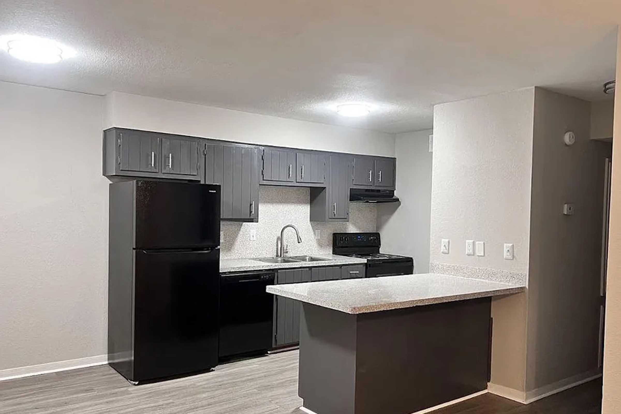 Modern kitchen featuring gray cabinets, black appliances including a refrigerator and stove, and a large countertop island. The space has neutral-colored walls, recessed lighting, and laminate flooring, creating a clean and contemporary look. Ideal for functional and stylish living.