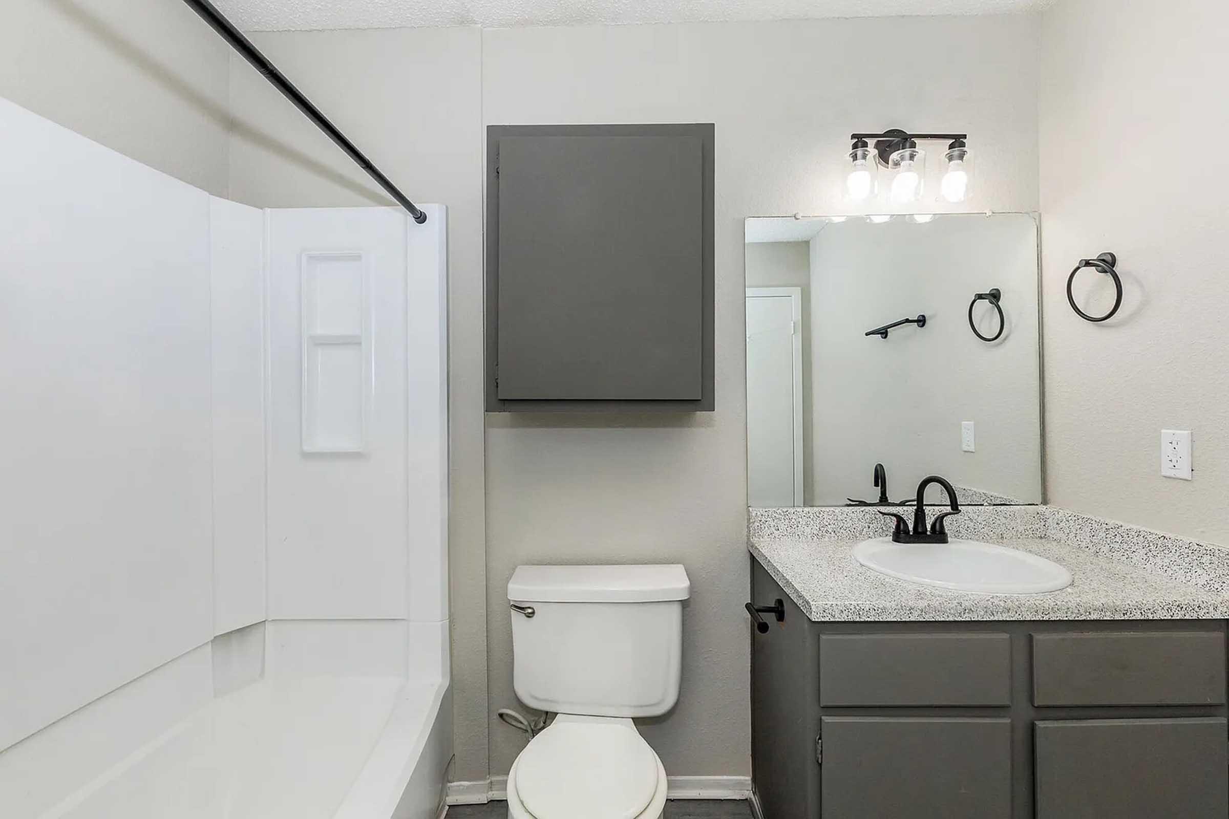 A modern bathroom featuring a bathtub with a shower, a wall-mounted mirror, a granite countertop sink, and a white toilet. The walls are painted in a neutral color, and there is a dark cabinet above the sink. Lighting is provided by a stylish fixture with multiple bulbs.