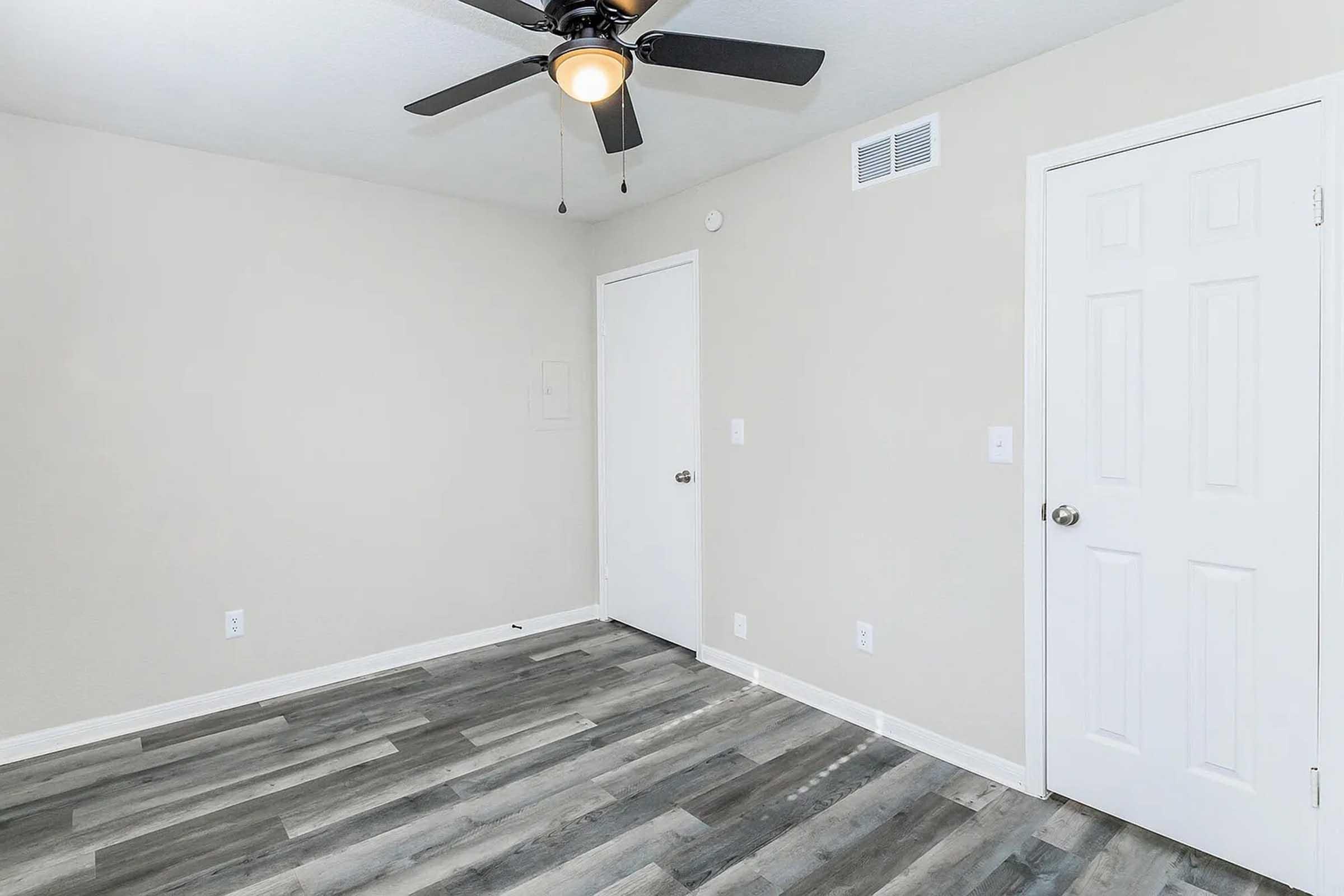 Empty room with light gray walls and a ceiling fan. Features a white door on the right, a door leading to another space, and light-colored wooden flooring. The space is bright and minimalistic, suggesting potential for customization or furnishing.
