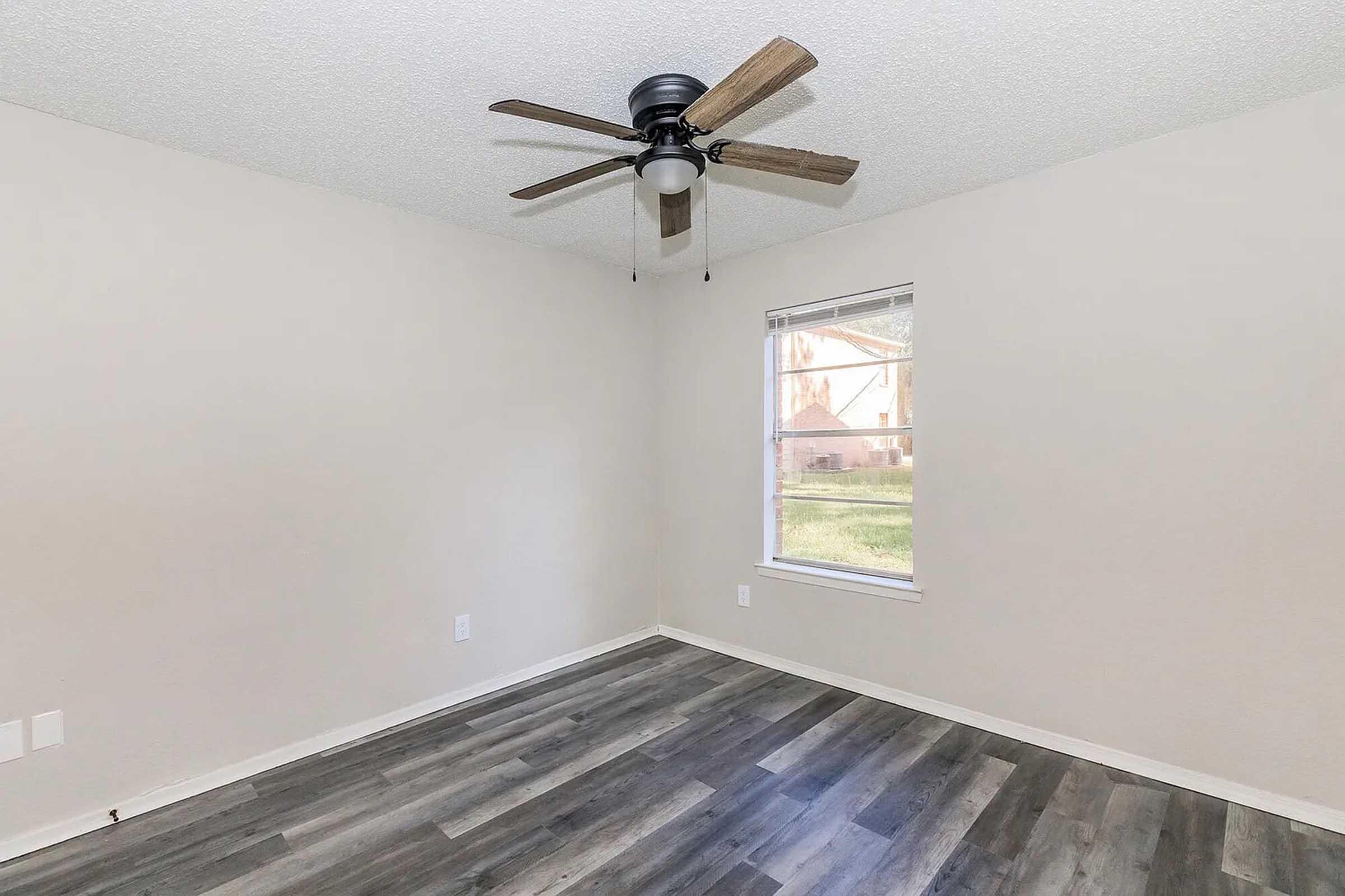 A spacious, empty room with light-colored walls and a ceiling fan. The floor features a modern, dark wood-like laminate. A window allows natural light to enter, showing an outdoor view.