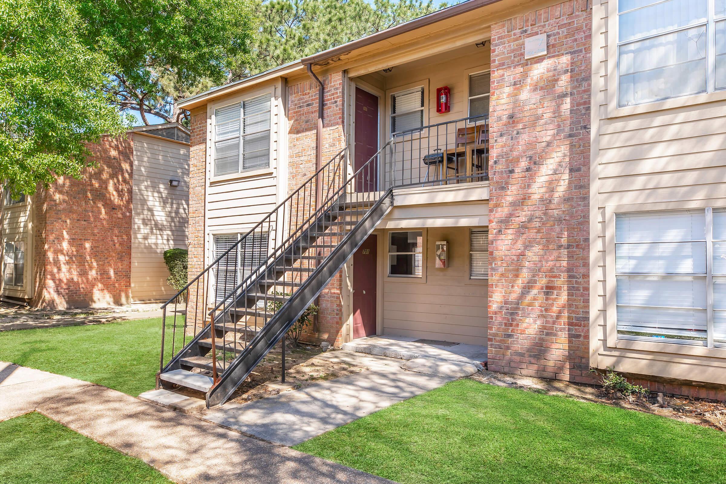 Exterior view of a two-story apartment building featuring a staircase leading to the upper level. The brick facade is complemented by greenery and a well-maintained lawn. The entrance of the upper apartment is visible, with a small balcony area.