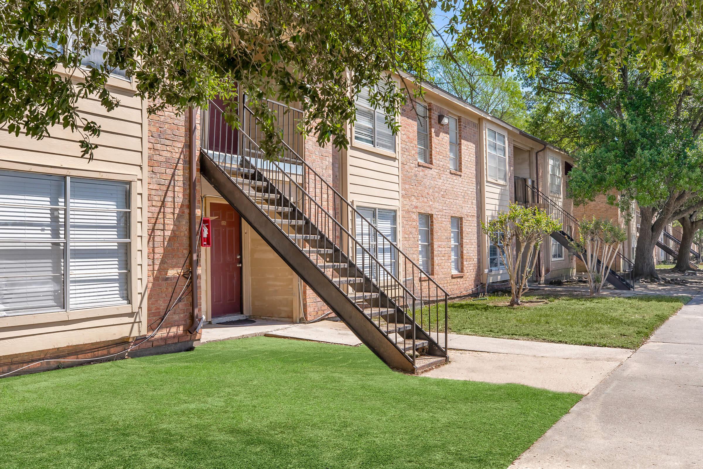 Exterior view of a two-story apartment building featuring a staircase leading to the upper floor, with large windows and a well-manicured lawn in the foreground. Trees provide shade, and the pathway is paved, creating a welcoming atmosphere.
