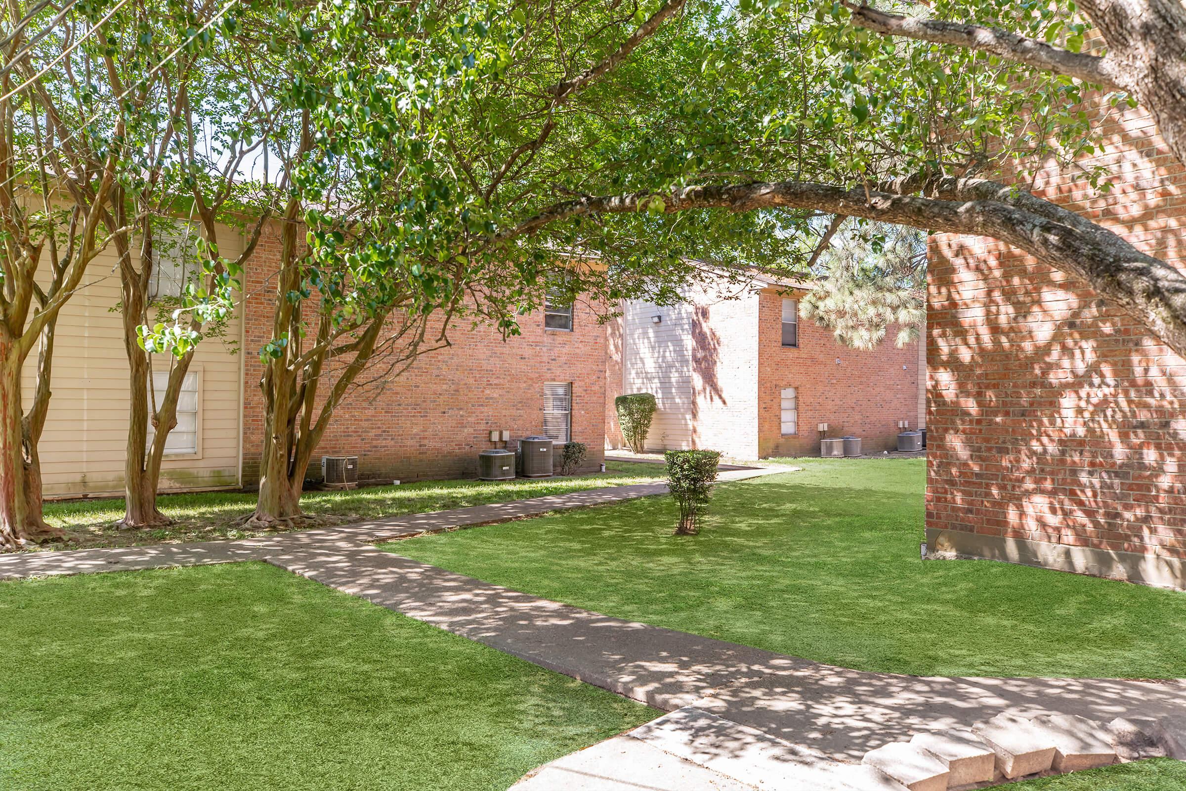 A landscaped outdoor area featuring green grass, trees, and several brick buildings. Pathways meander between the structures, with air conditioning units visible along the walls, creating a serene environment. Sunlight filters through the leaves, casting shadows on the ground.