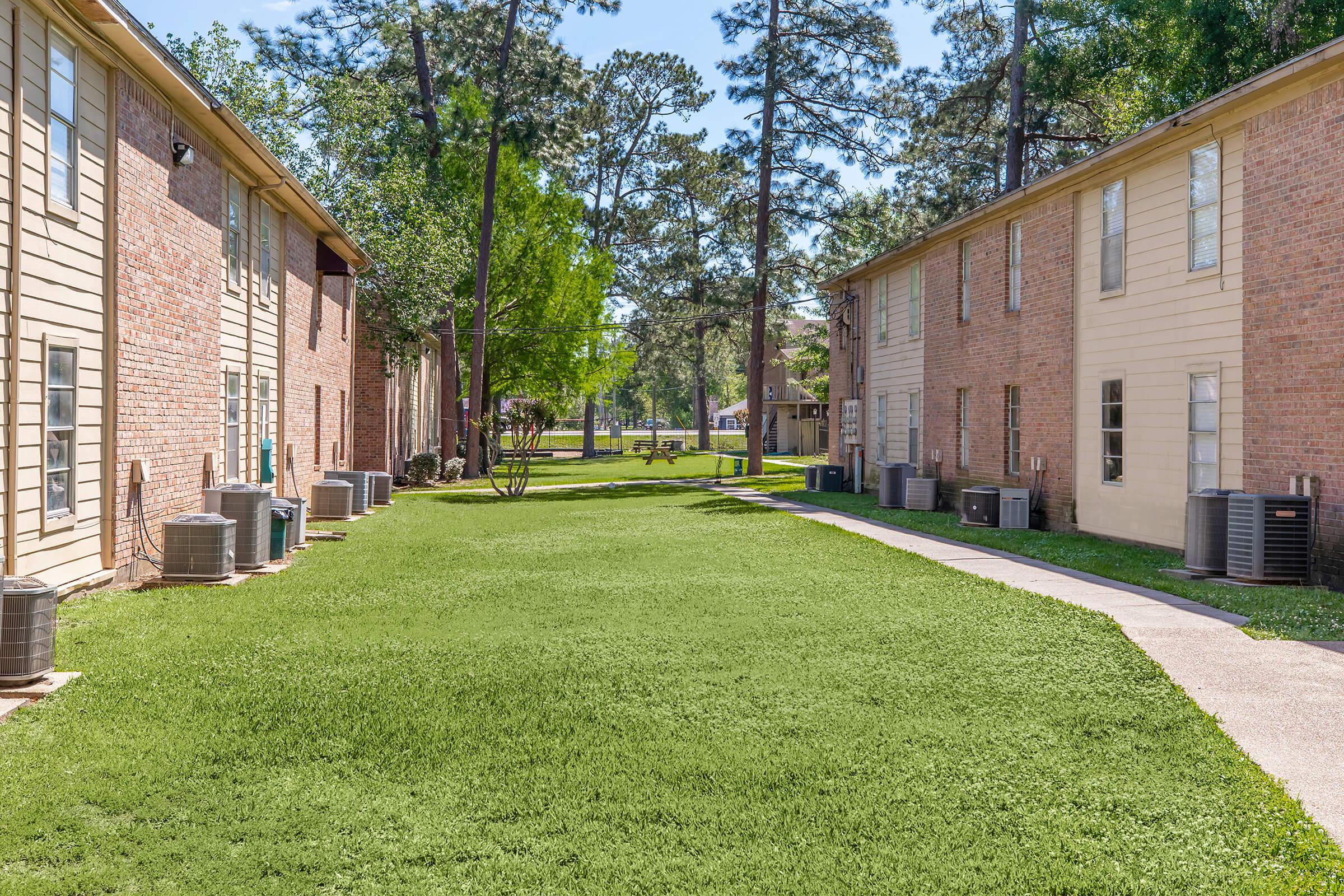 A well-maintained green lawn between two rows of beige and brick apartment buildings, with trees and shrubs in the background. Air conditioning units are placed along the sides of the buildings, and a clear blue sky is visible above.