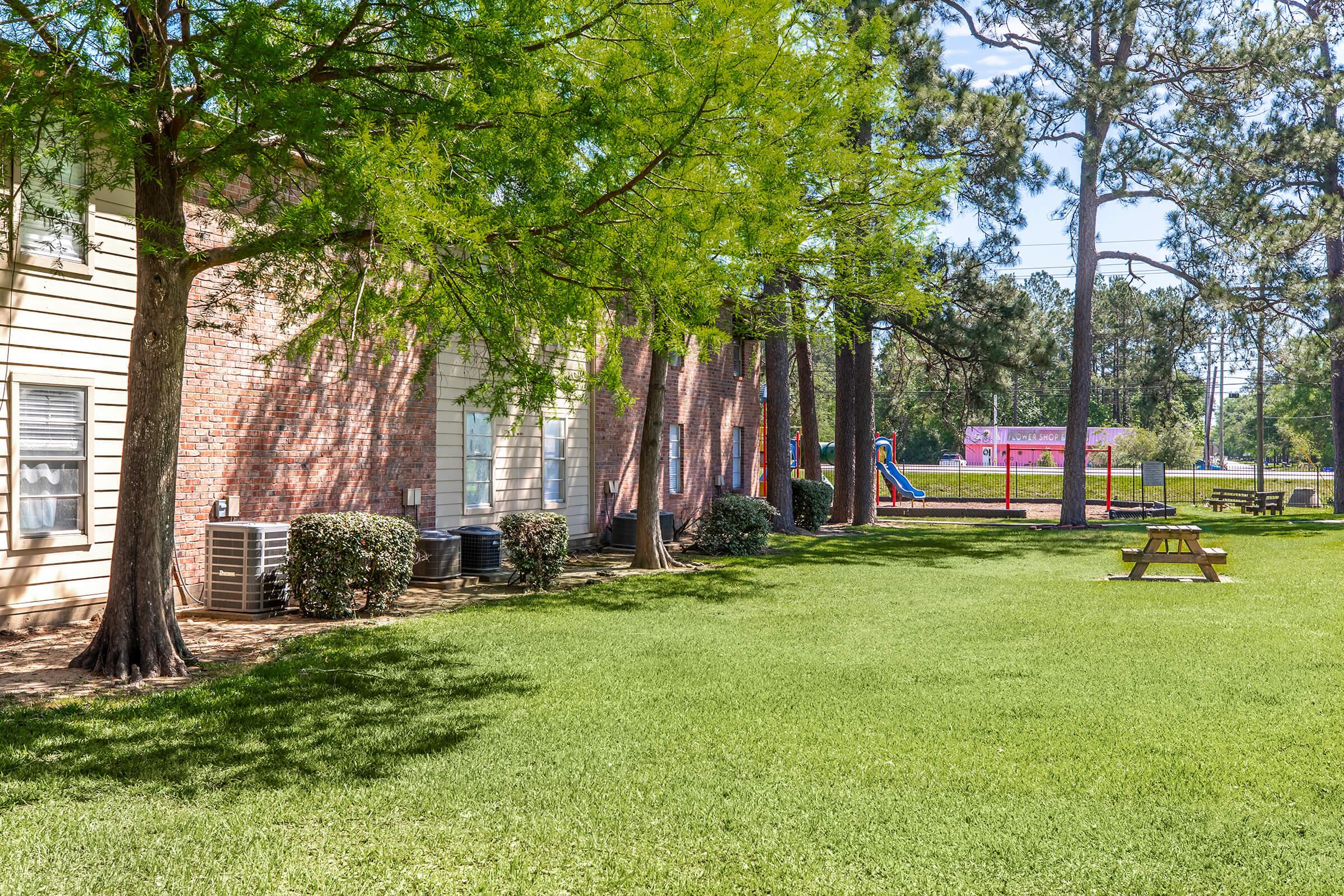 A peaceful outdoor scene featuring a well-maintained lawn with lush green grass, a picnic table, and trees providing shade. In the background, there are brick apartment buildings and a playground with colorful equipment. The setting is bright and inviting, perfect for relaxation and outdoor activities.