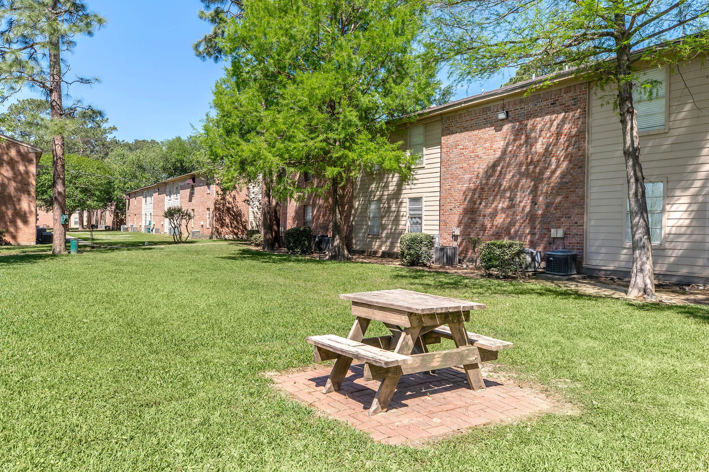 A grassy area featuring a wooden picnic table surrounded by trees and brick-apartment buildings. The scene is sunny and well-maintained, ideal for outdoor relaxation or gatherings.