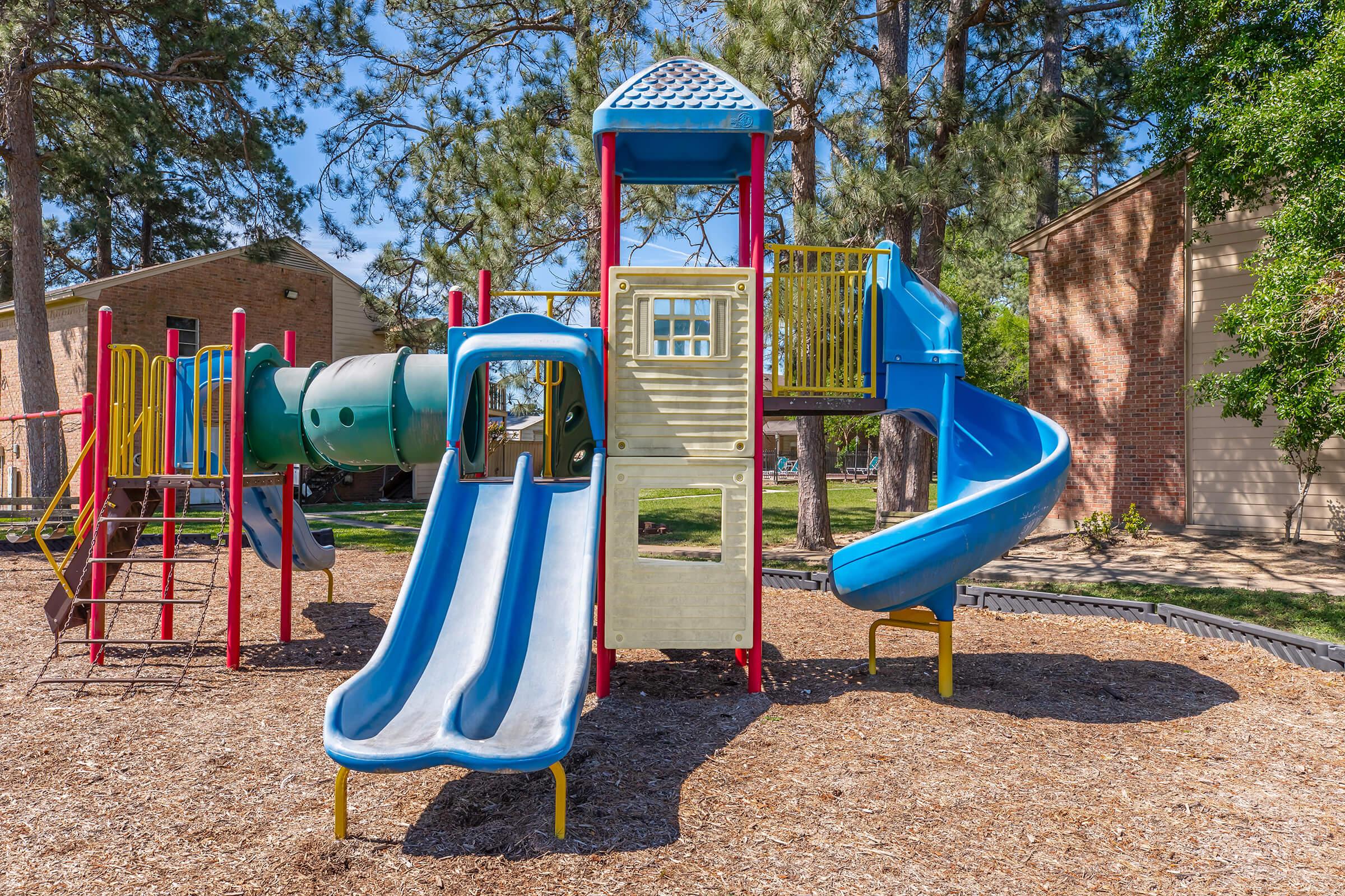 Colorful playground structure featuring blue and yellow slides, a climbing frame, and a tubular slide. The equipment is set on a soft, mulch surface, surrounded by trees and adjacent to brick buildings, creating a fun outdoor play area for children.