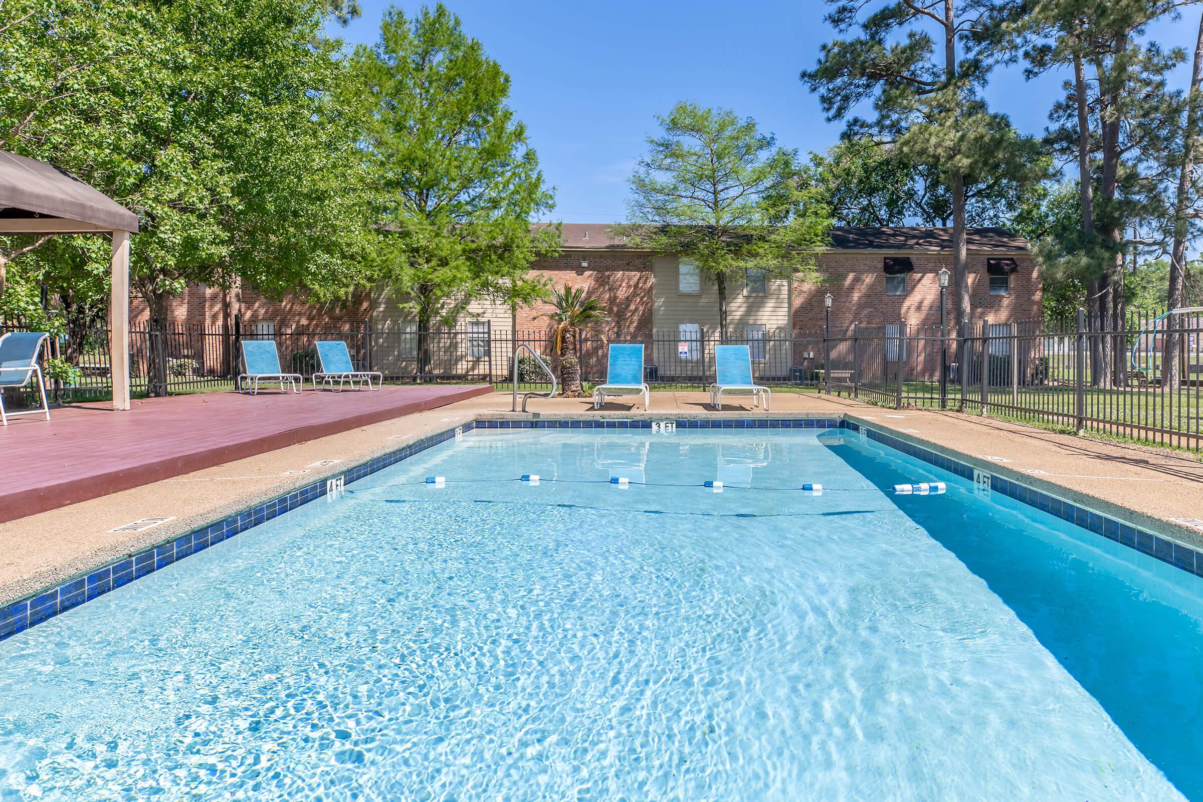 A clear blue swimming pool surrounded by lounge chairs and a shaded gazebo. The pool is partially filled with water, and lush green trees and a brick building can be seen in the background under a bright blue sky. The area is well-maintained and inviting for relaxation and leisure activities.