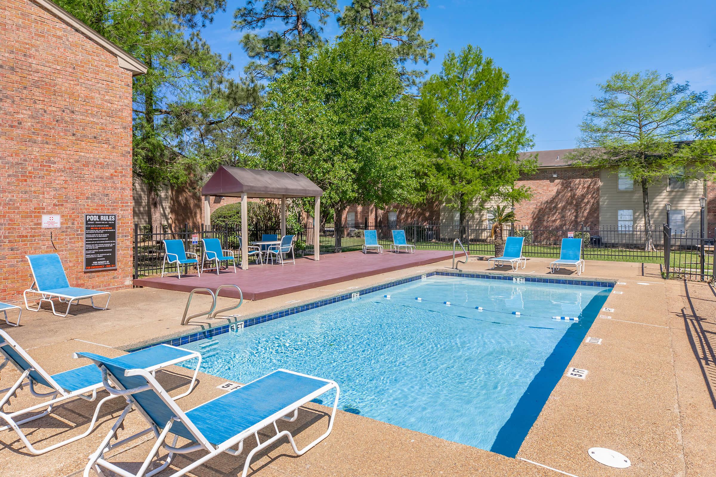 A clean outdoor pool area featuring blue lounge chairs around the pool, a shaded seating area, and surrounded by greenery. The pool has clear water and is sectioned off with a safety fence. Bright blue sky above adds to the inviting atmosphere.