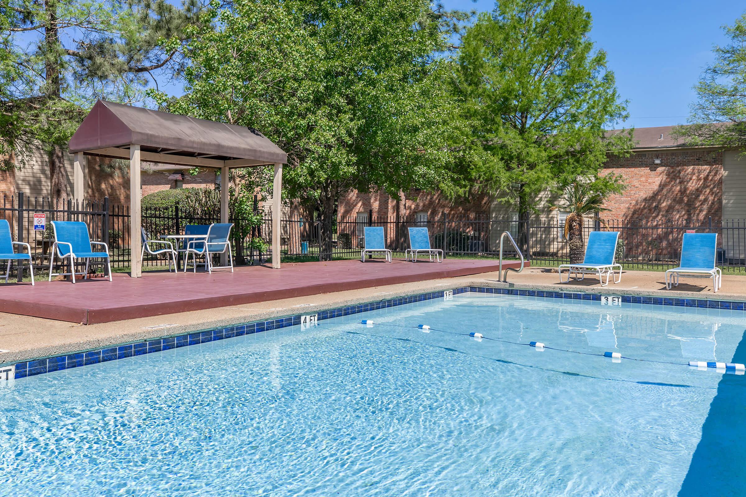 A clear swimming pool surrounded by a fence, with lounge chairs and a shaded gazebo nearby. Lush green trees provide shade, and the sky is bright and blue, creating a relaxing outdoor atmosphere.