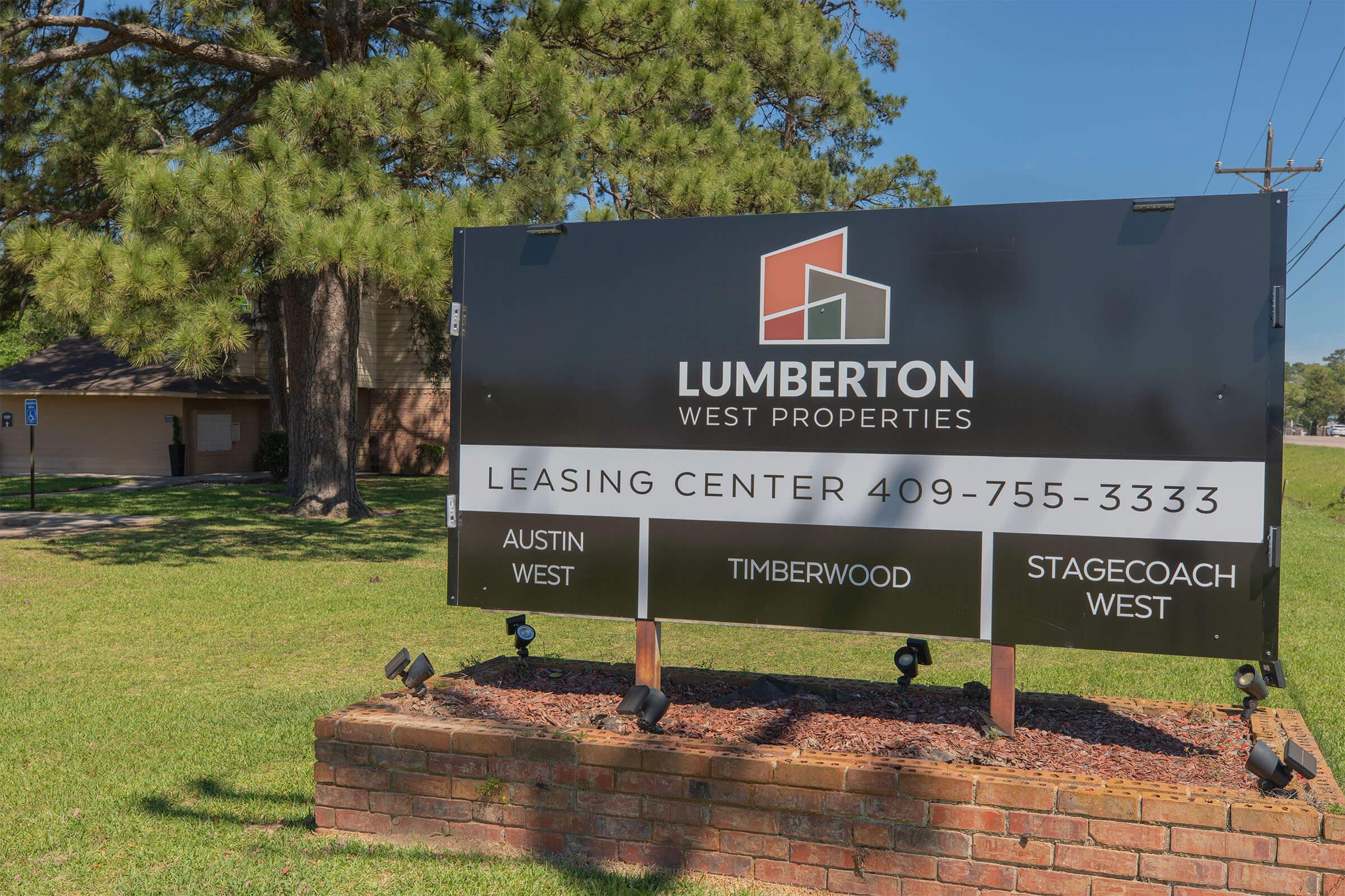Sign for Lumberton West Properties, featuring a leasing center with contact number 409-755-3333. The sign lists three property names: Austin West, Timberwood, and Stagecoach West. The background includes trees and a nearby building, set in a grassy area.