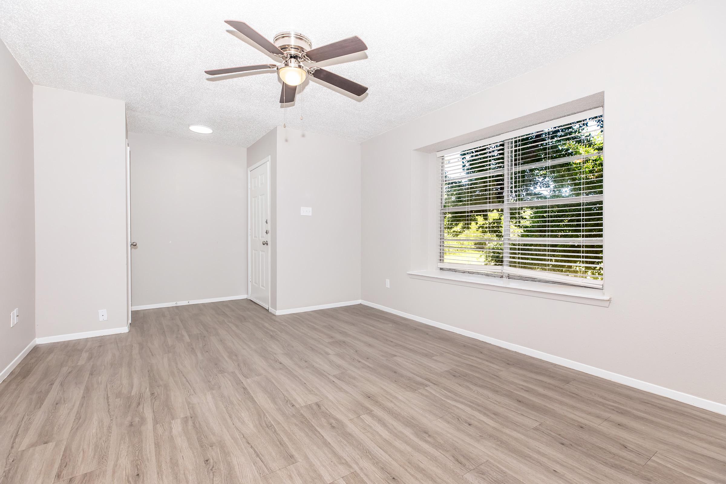 Empty room with light gray walls and new wood-like flooring. There is a ceiling fan with four blades and a large window with white blinds letting in natural light. A door is visible on the left side, and the space is unobstructed, creating an open, inviting atmosphere.