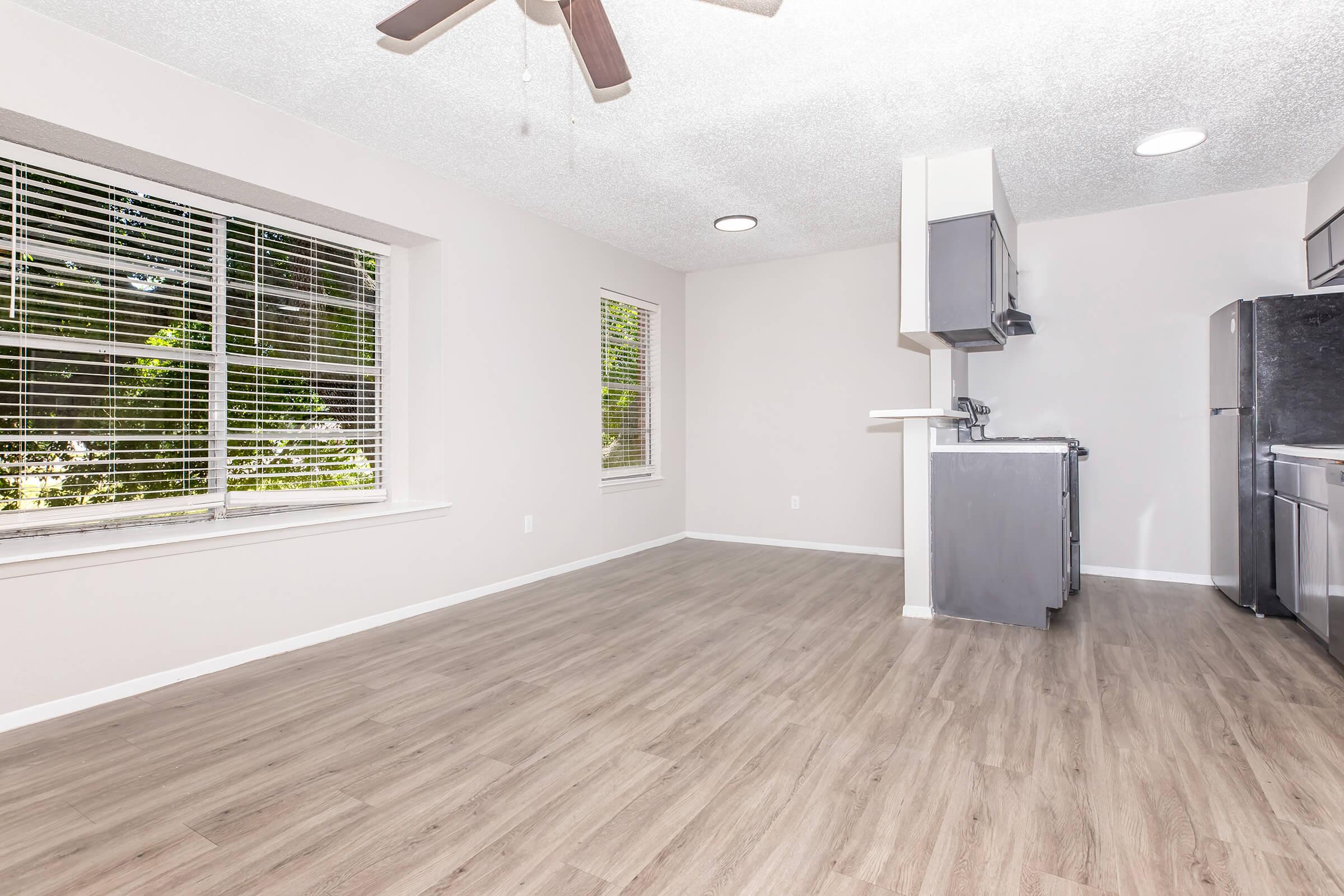 Interior view of a modern, well-lit apartment featuring a spacious living area with large windows, light-colored walls, and hardwood-style flooring. A ceiling fan hangs above, and there is a kitchen area on one side with gray cabinetry and stainless steel appliances.