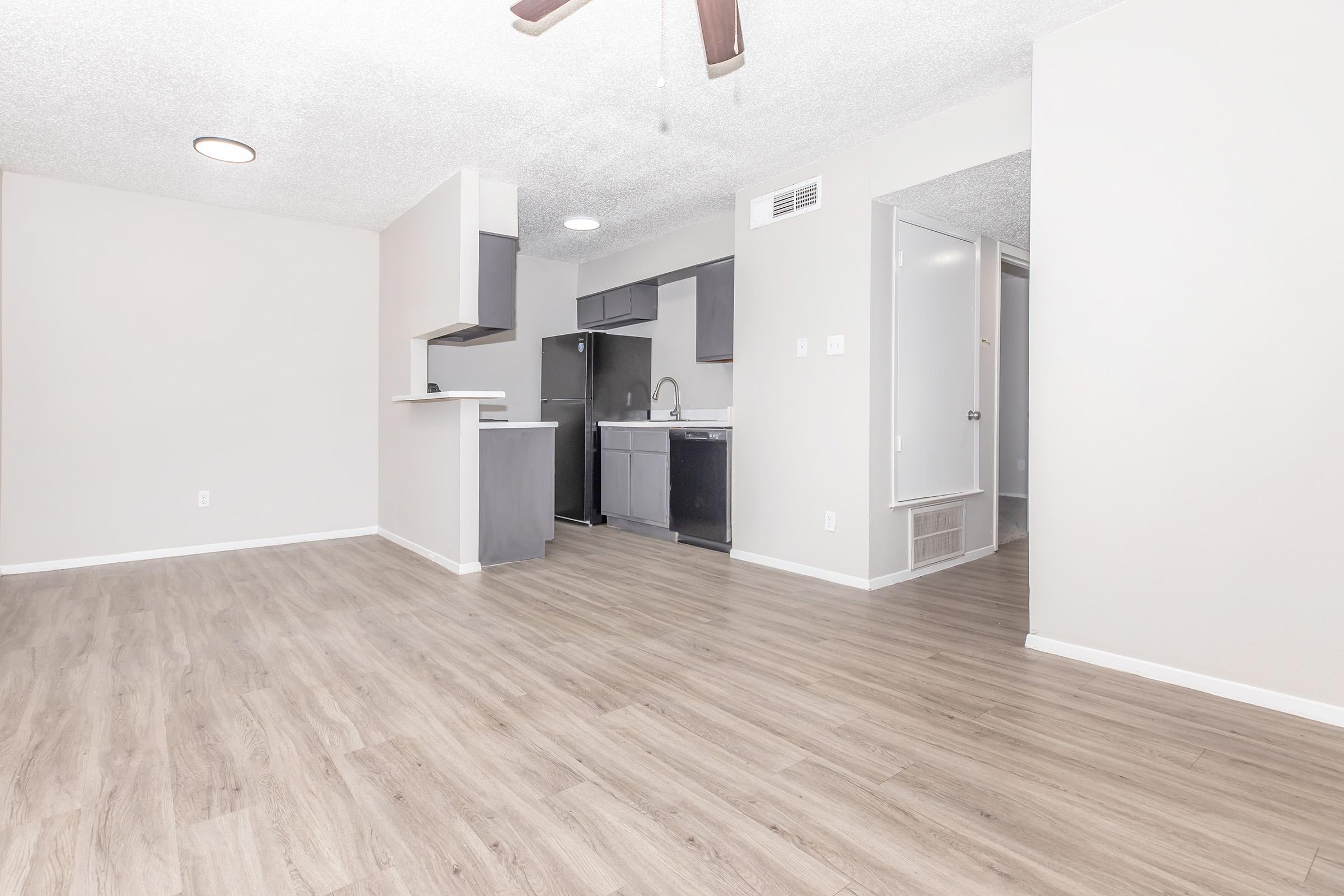 Spacious interior of a modern apartment, featuring light-colored walls and hardwood floors. The kitchen area is visible with dark cabinetry and appliances, while a ceiling fan hangs above. A doorway in the background suggests access to another room or bathroom, creating an open and inviting living space.