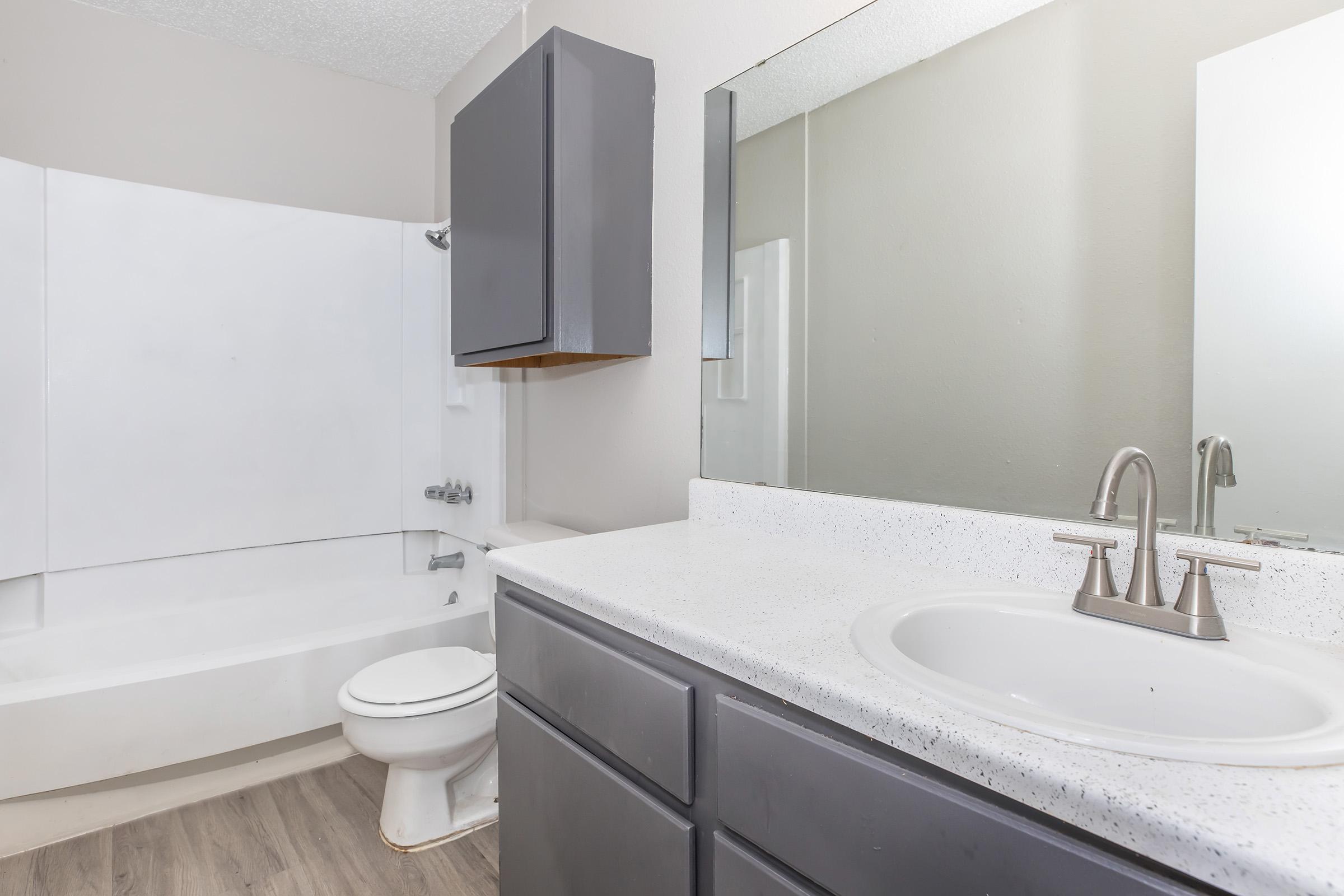 A clean and modern bathroom featuring a white bathtub, a gray cabinet above the toilet, and a sink with a polished chrome faucet. The counter is made of light-colored stone, and there is a large mirror above the sink, with neutral-colored walls and flooring.