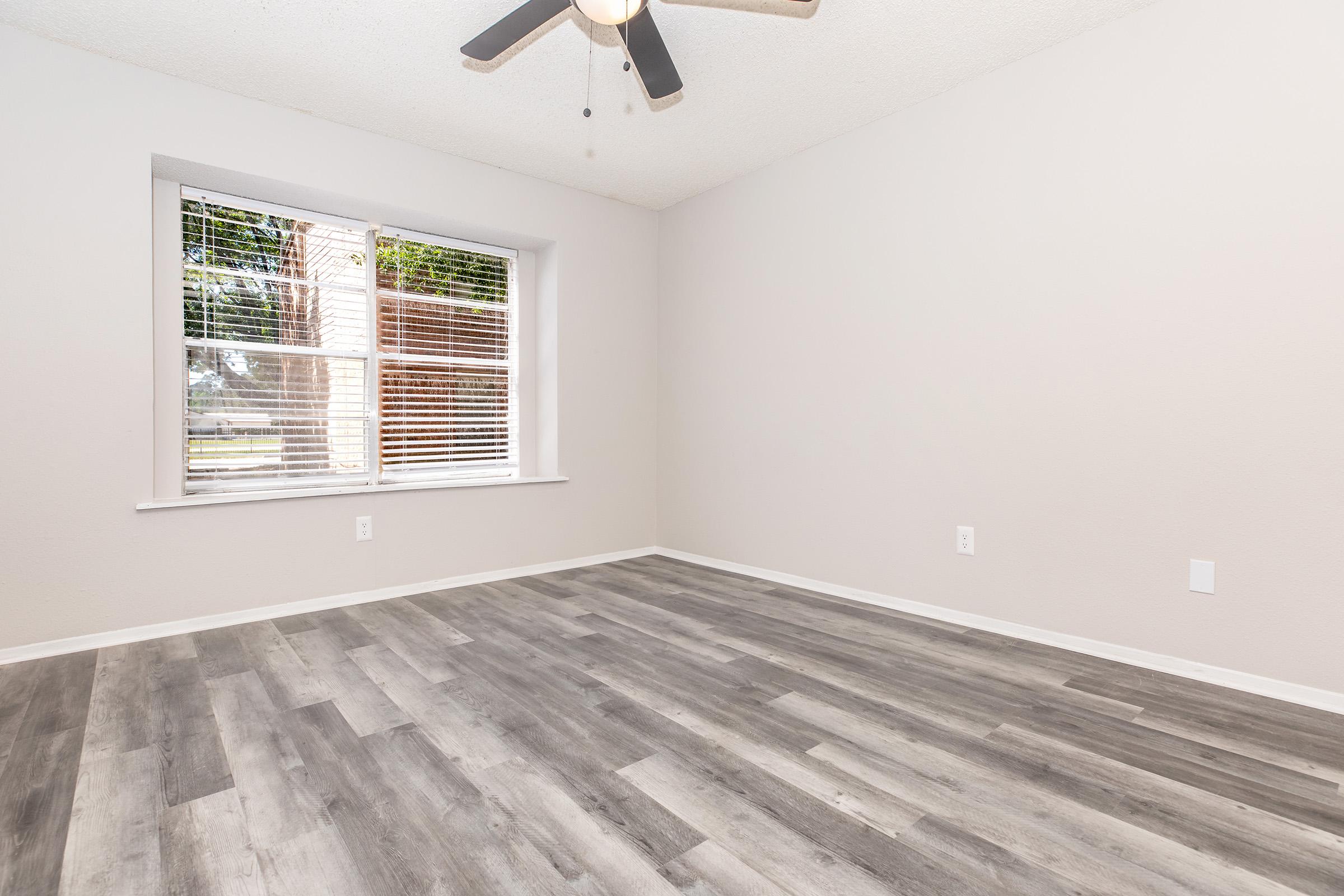 A view of an empty room with light gray walls and a ceiling fan. The floor features laminate planks in a gray tone. A window with blinds lets in natural light, showcasing a pleasant exterior view of greenery. The space is well-lit and ready for decoration or furniture.