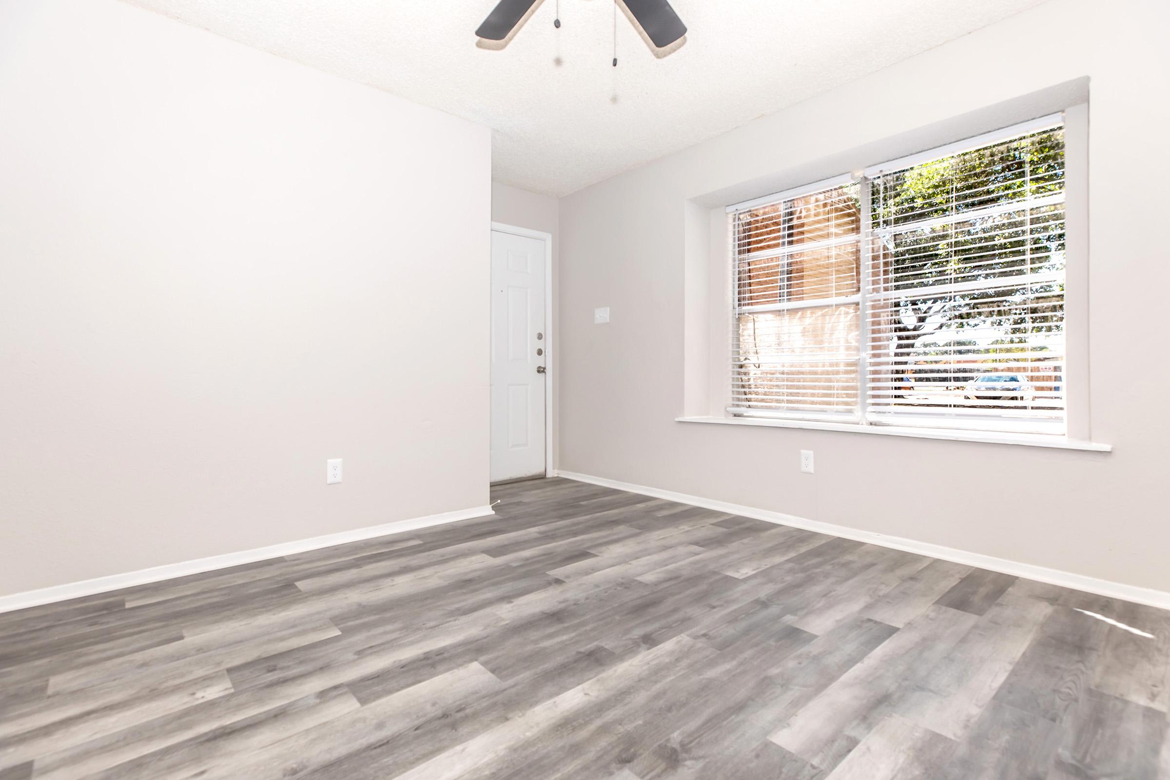 A well-lit, empty room featuring light gray walls and new laminate flooring. A ceiling fan is positioned above, and a window with blinds allows natural light to enter. The entrance door is visible, providing access to the outside. The overall space is clean and ready for furniture placement.