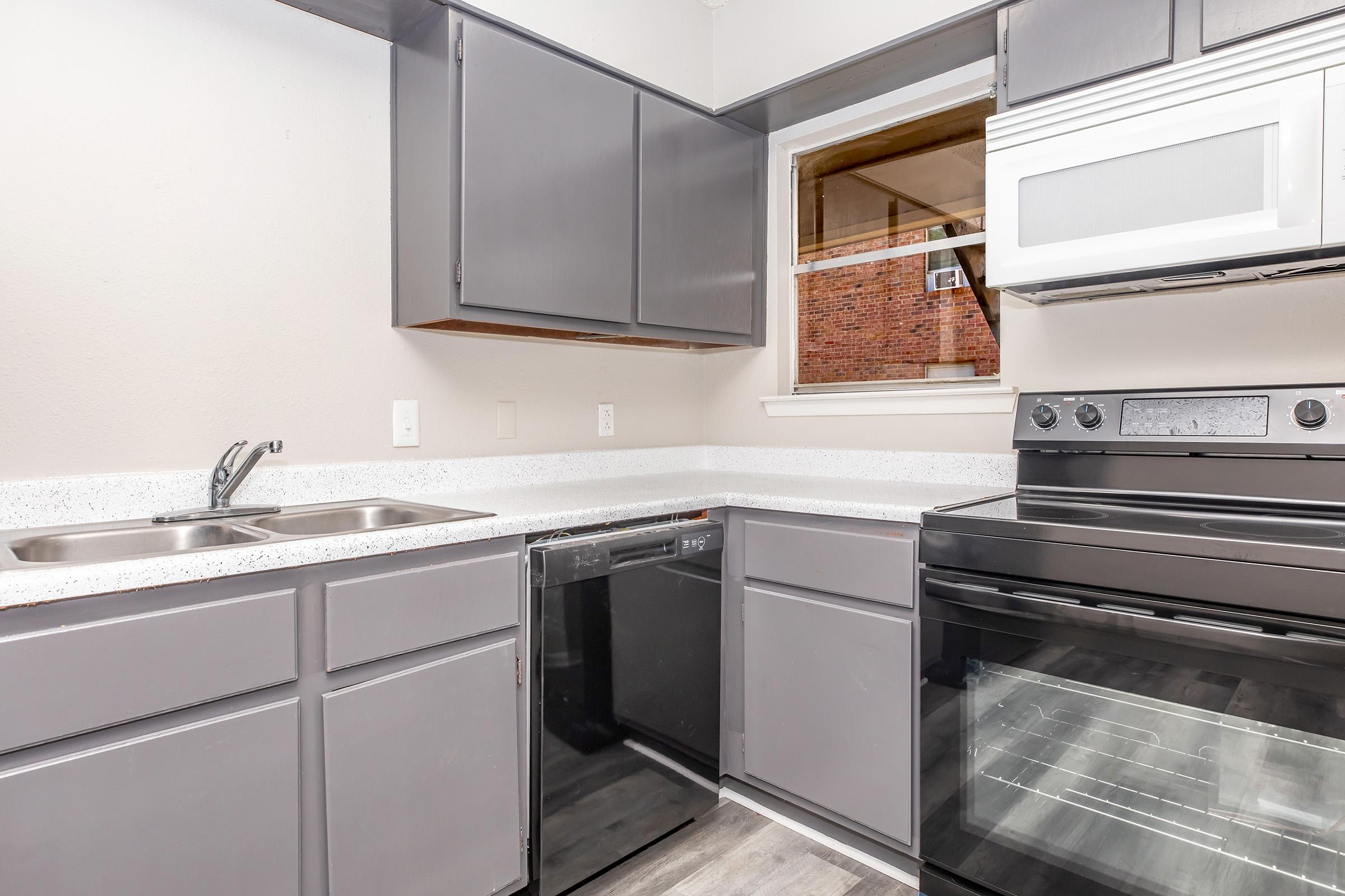 A modern kitchen featuring gray cabinets, a stainless steel sink, a dishwasher, and a black stove. There is a window above the sink, allowing natural light to brighten the space. The countertops are a light color, contrasting with the darker cabinetry, creating a sleek and contemporary look.